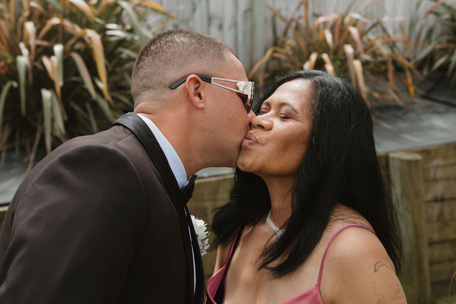 A couple shares a romantic kiss while standing outdoors in formal attire.