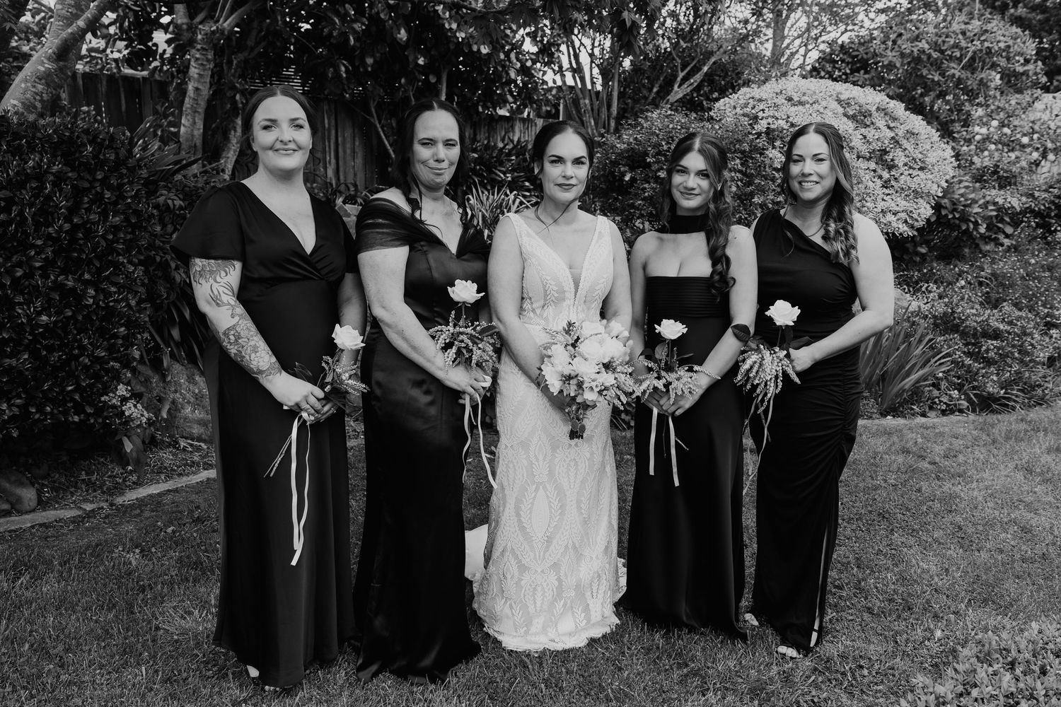 A wedding party poses outdoors in black and white formal attire with bridesmaids holding bouquets.