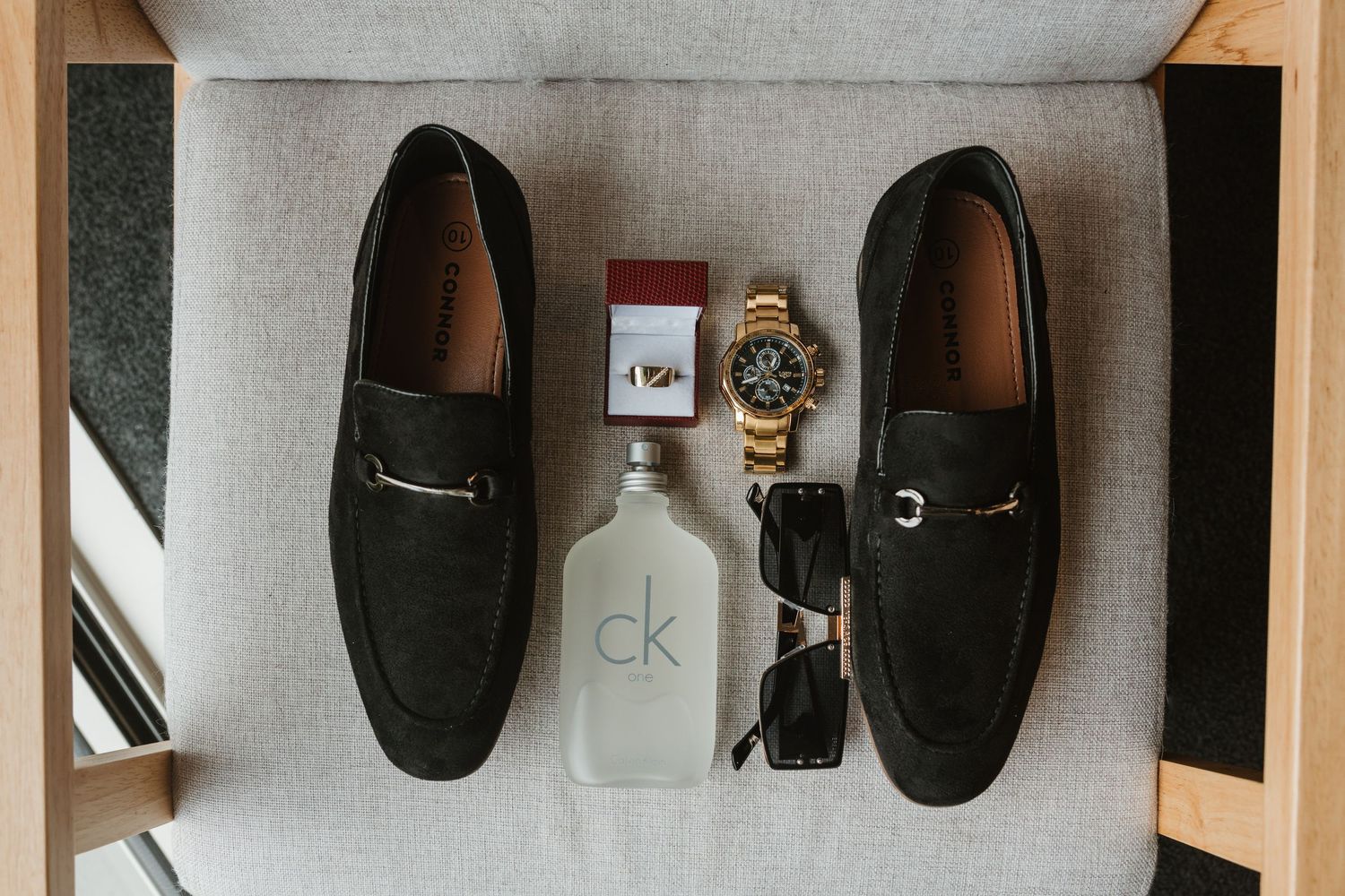 Luxury men's accessories flatlay with black suede loafers, CK vodka bottle, gold watch and sunglasses on gray surface.