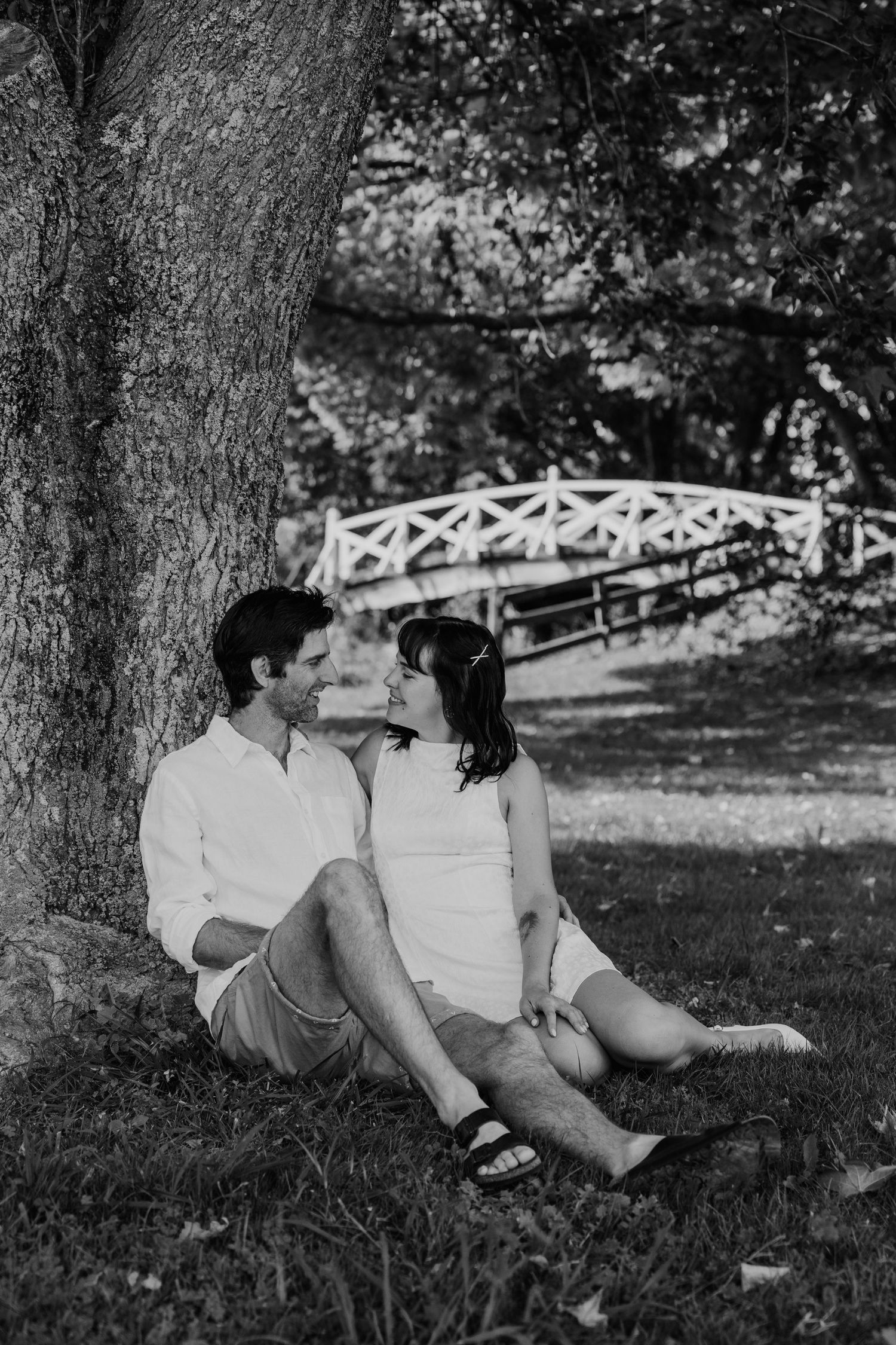 Black and white photo of a couple sitting together under a large tree in a park with a white bridge in the background.