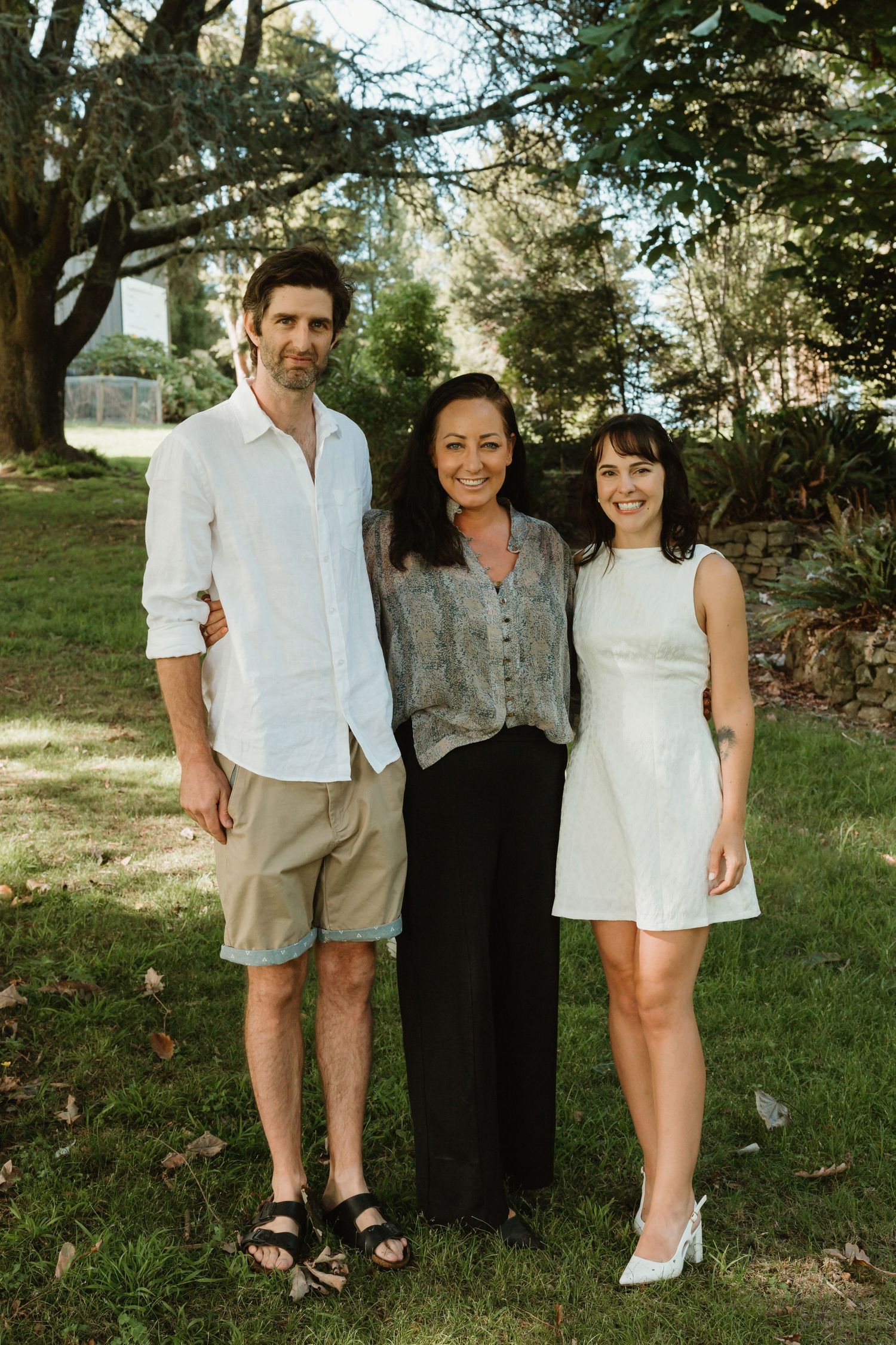 Three people smiling and posing together outdoors in a lush green garden setting on a sunny day.