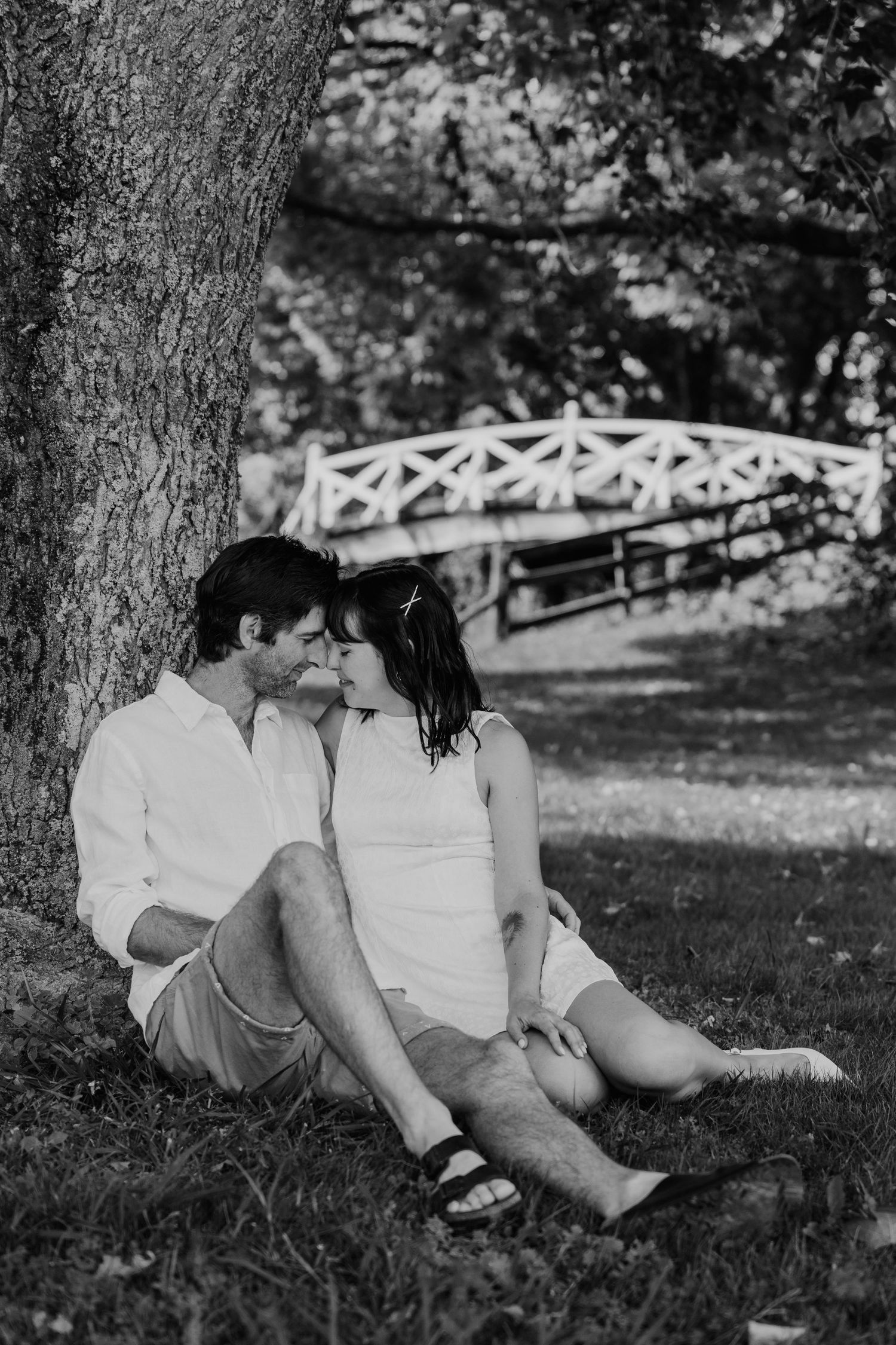Black and white photo of a romantic couple sitting together under a tree in a park with a white bridge in the background.