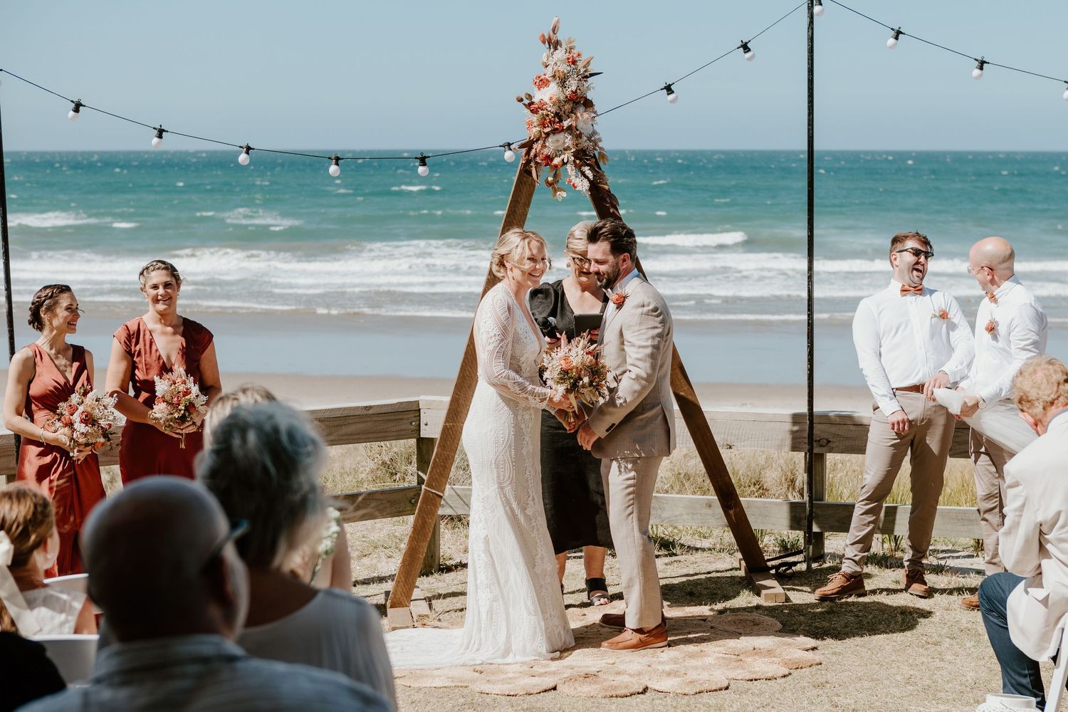 Panoramic view of a beach wedding ceremony with guests seated under string lights by the ocean. Papamoa Surf Lifesaving Club Wedding