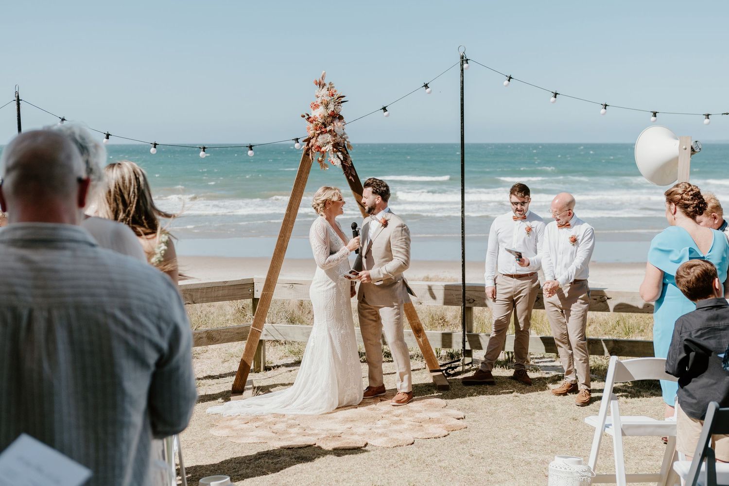 A wedding ceremony setup on a beach with string lights and an ocean view backdrop. Papamoa Surf Lifesaving Club Wedding