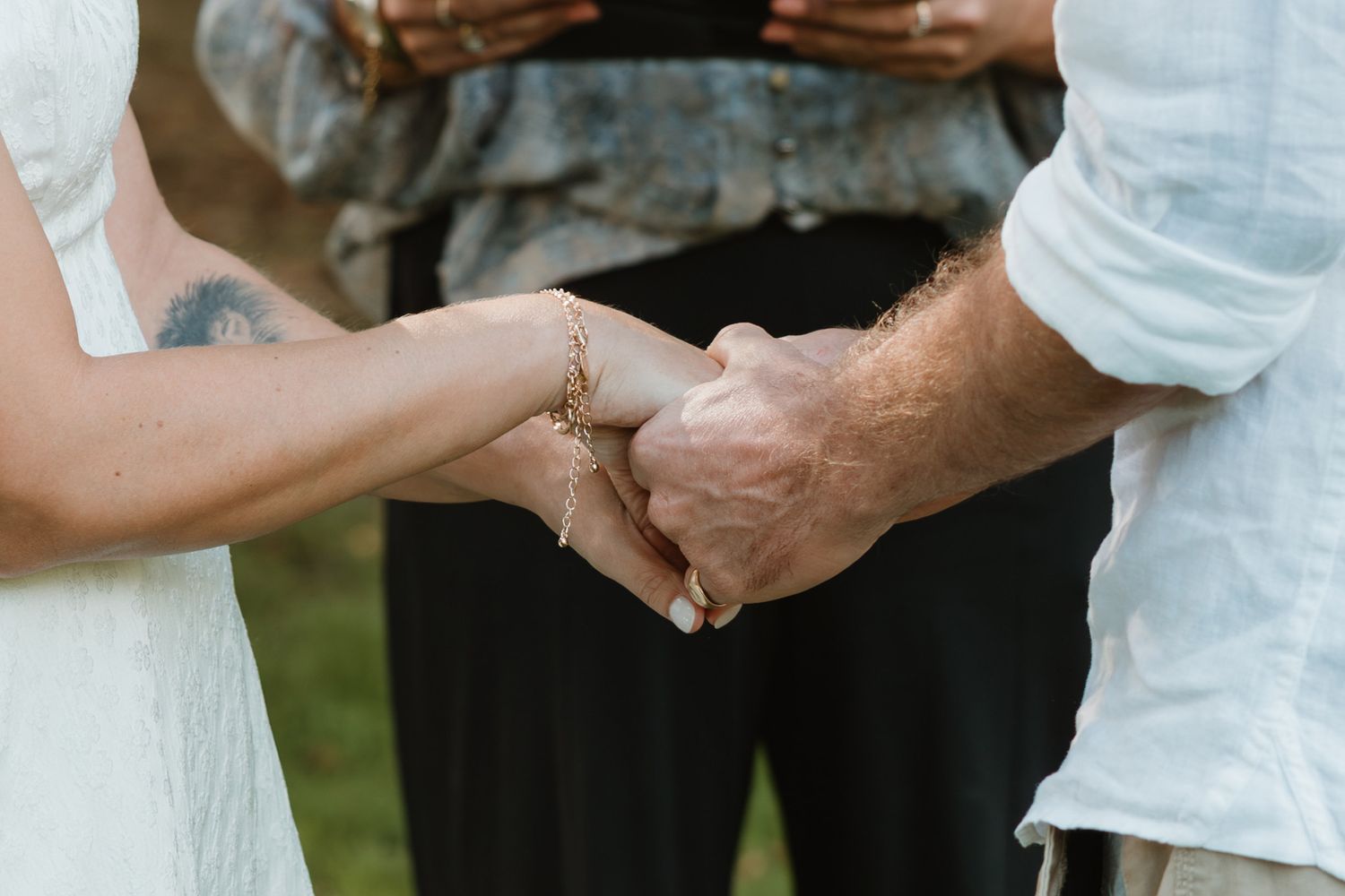 Couple holding hands during wedding ceremony with officiant in background, bride showing floral tattoo on wrist.
