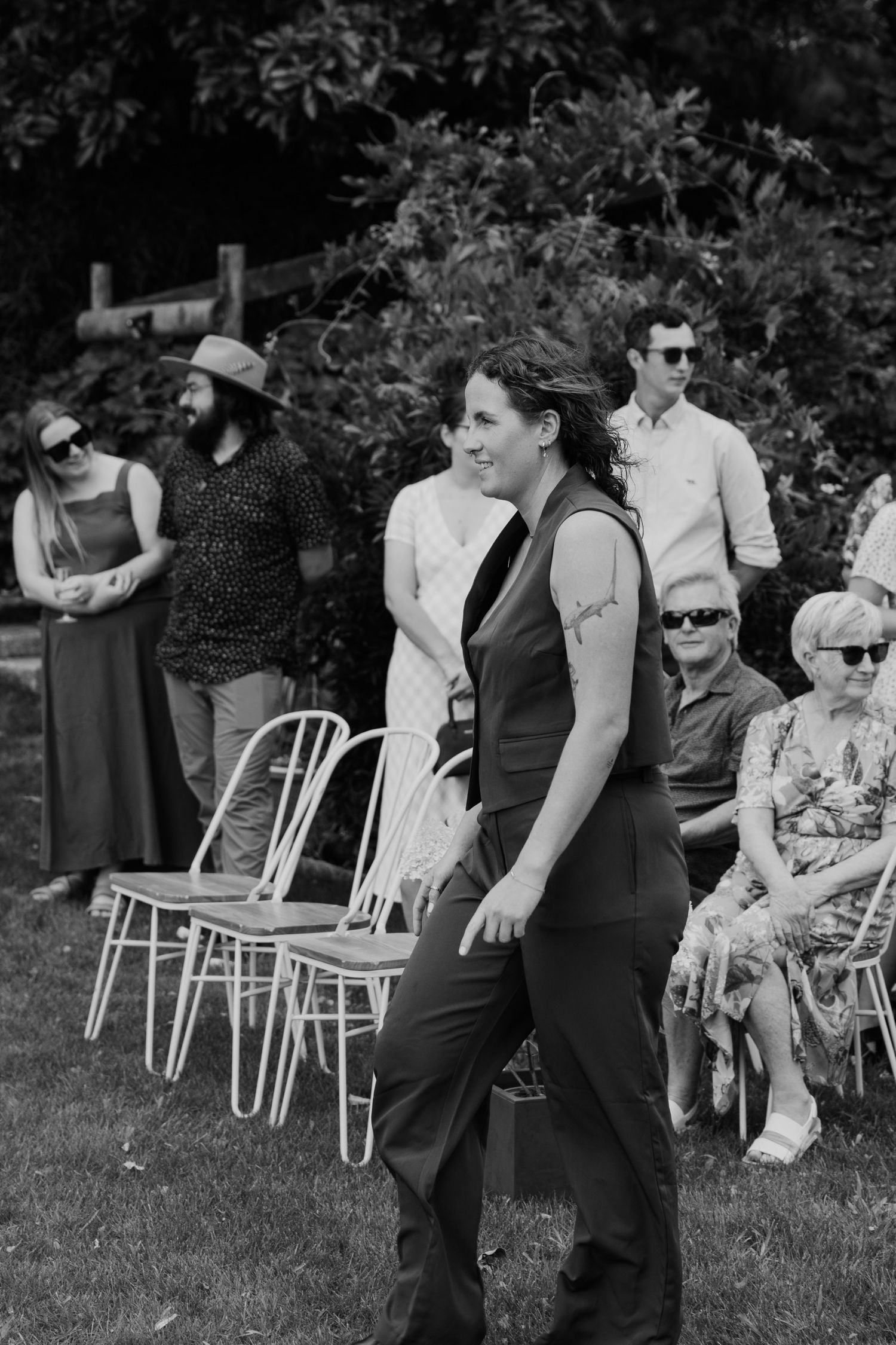 Black and white outdoor wedding scene with guests seated and standing on grass, woman in sleeveless jumpsuit smiling in foreground.