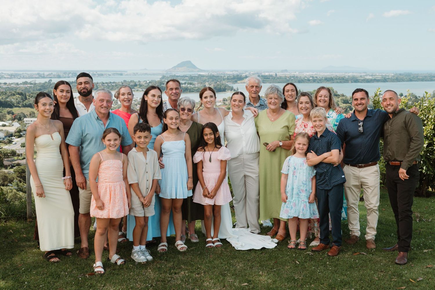Large family group posing outdoors with scenic ocean and mountain views in the background on a sunny day.