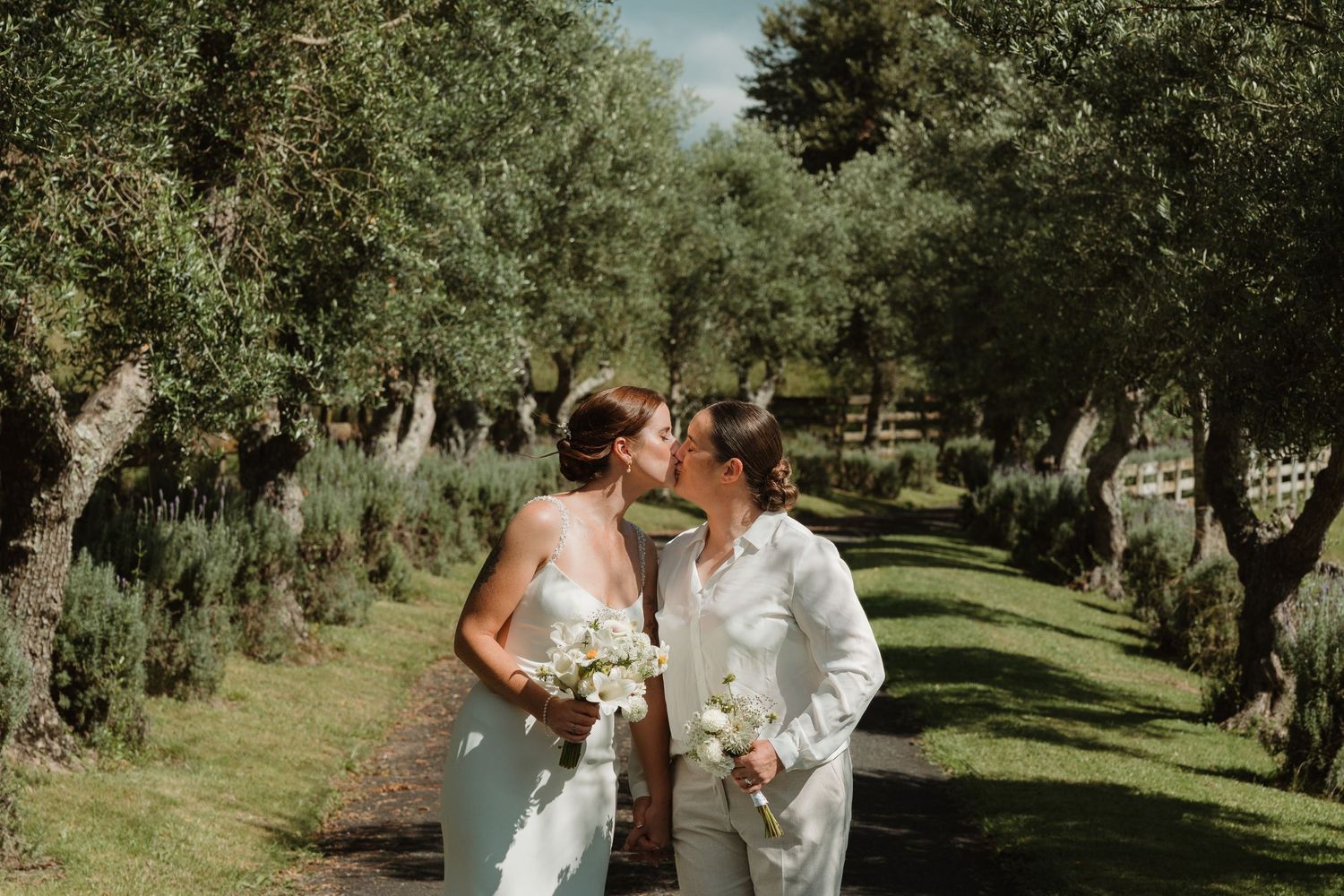 Two brides sharing a kiss on a tree-lined path, holding white bouquets on their wedding day.