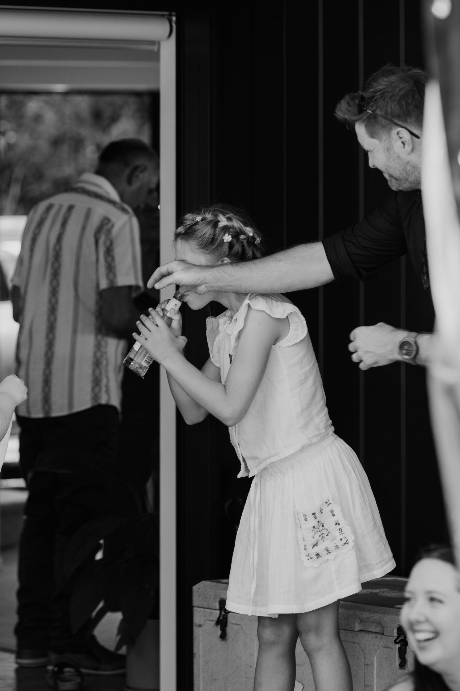Black and white photo of a girl in white dress receiving help from a man opening a bottle at an outdoor event.