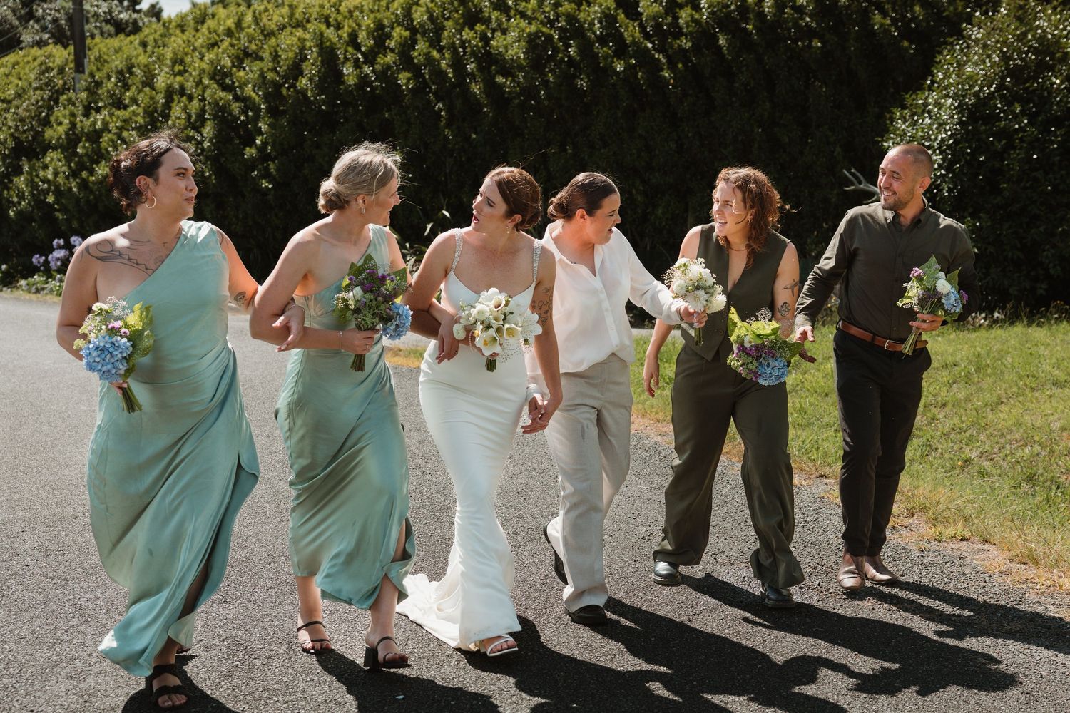 Bride and bridesmaids in mint green dresses walking together outdoors, holding bouquets on a sunny wedding day.