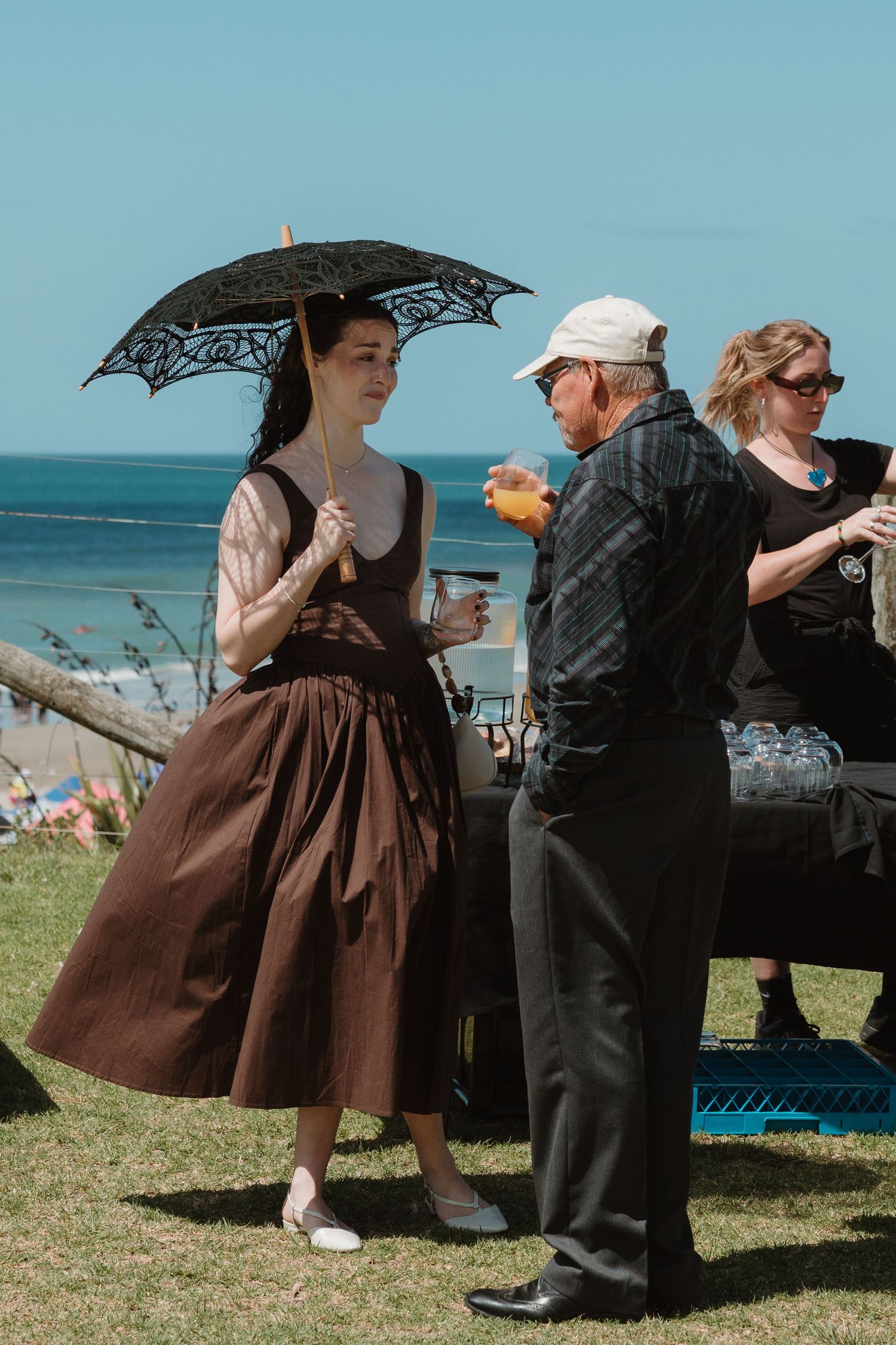 Woman in vintage brown dress holding lace parasol at outdoor seaside event on sunny day.