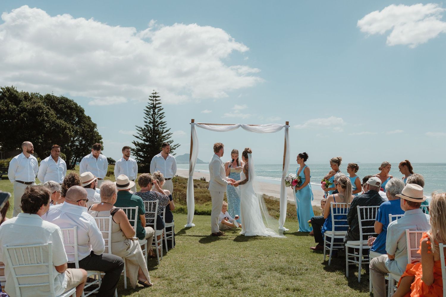 Outdoor beach wedding ceremony with guests seated on white chairs as couple exchanges vows under a floral arch by the ocean.