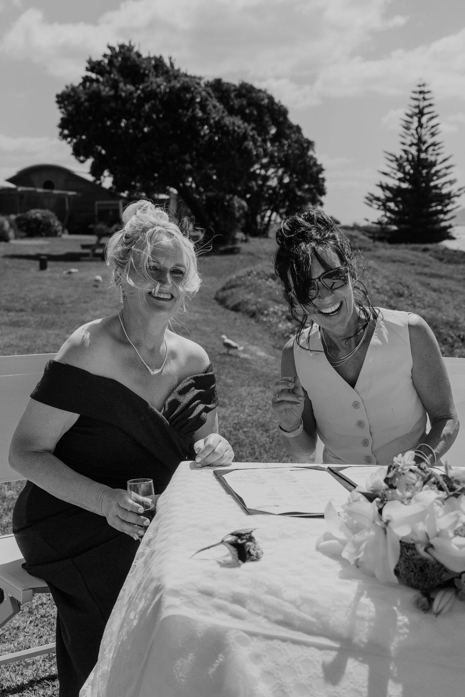 Two women laughing together at an outdoor wedding signing table, black and white photo.