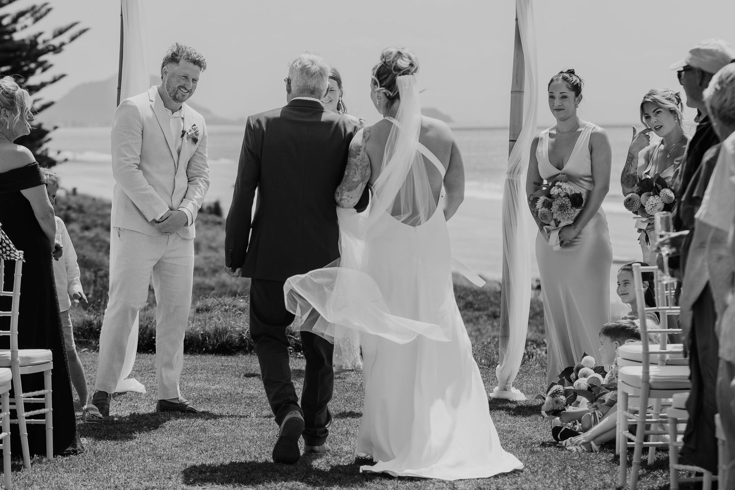 Black and white outdoor wedding ceremony with bride in backless gown being walked down the aisle by her father.