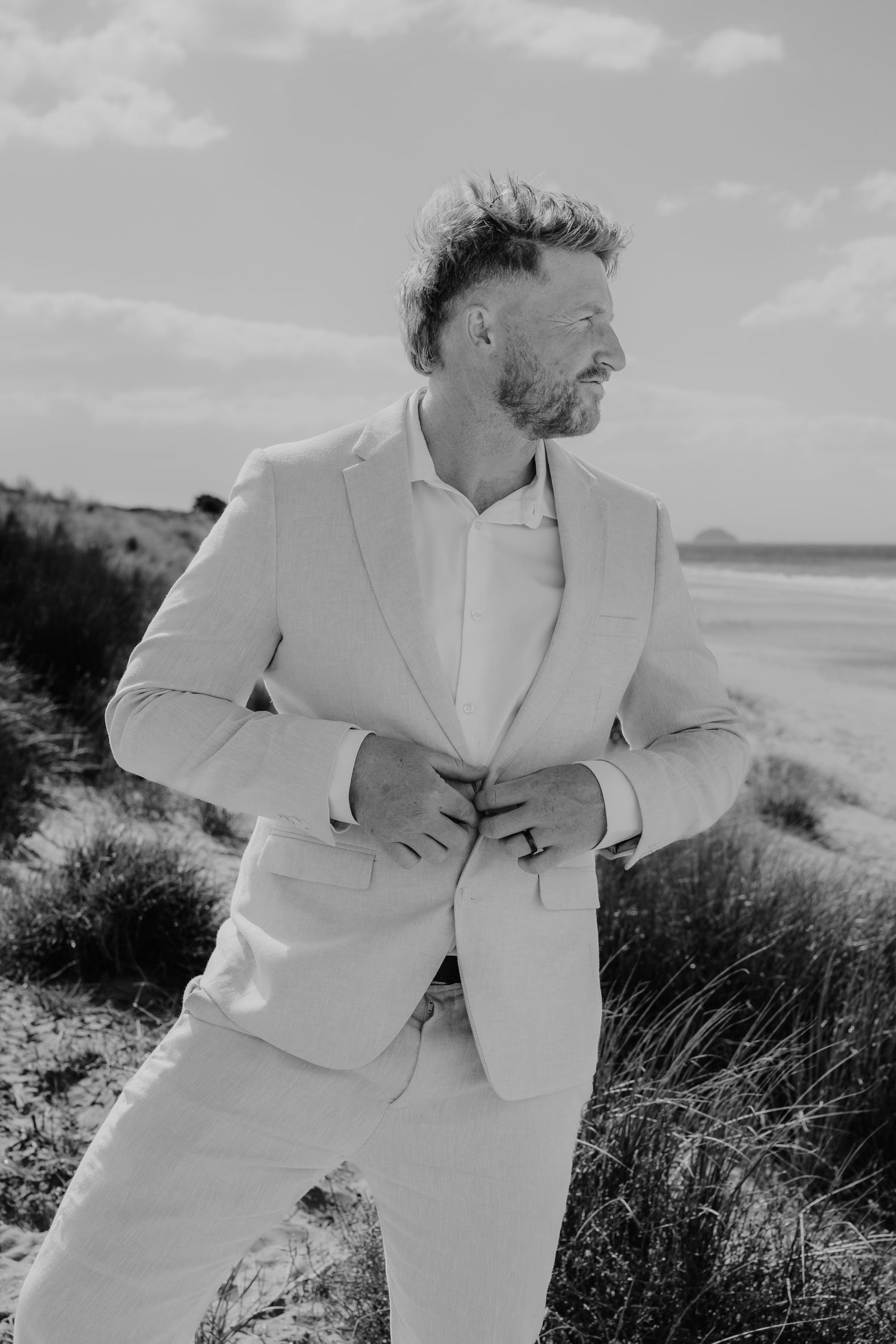 Black and white photo of a well-dressed man in a light suit buttoning his jacket on a sandy beach.