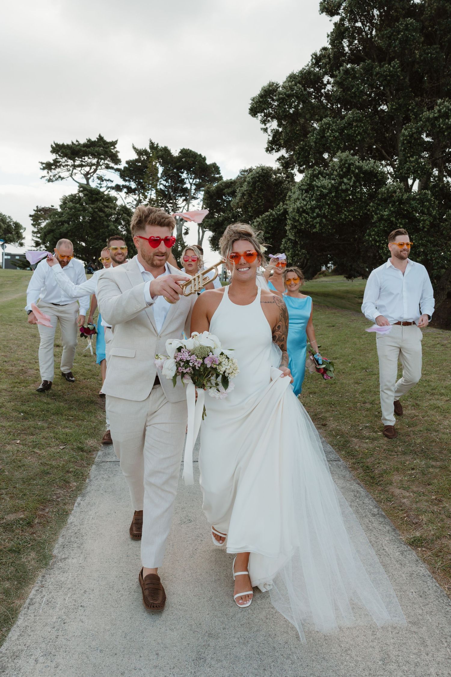 Bride and groom in matching red sunglasses lead wedding party down path, bride holding bouquet, groom with champagne.