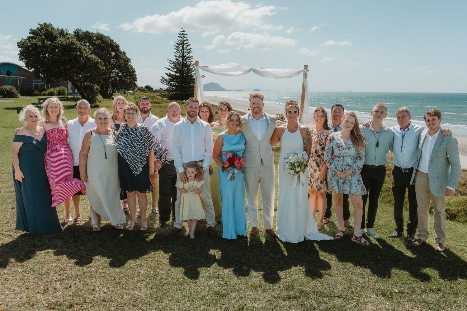 Wedding party posing outdoors near ocean altar on sunny day, bride in white, guests in formal attire on green lawn.