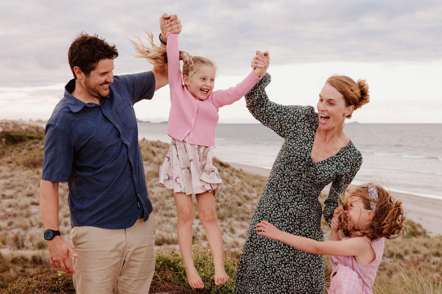 A happy family spends time together lifting children up while playing on a cloudy beach. Tauranga lifestyle family photographer