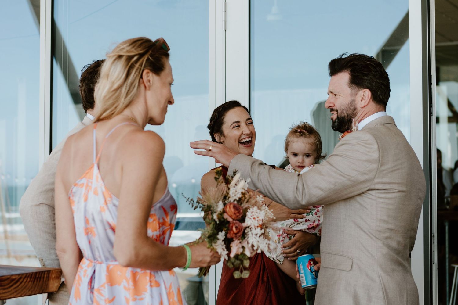 Wedding party celebrates joyfully with laughter and hugs by large glass windows overlooking the ocean. Papamoa Surf Lifesaving Club Wedding