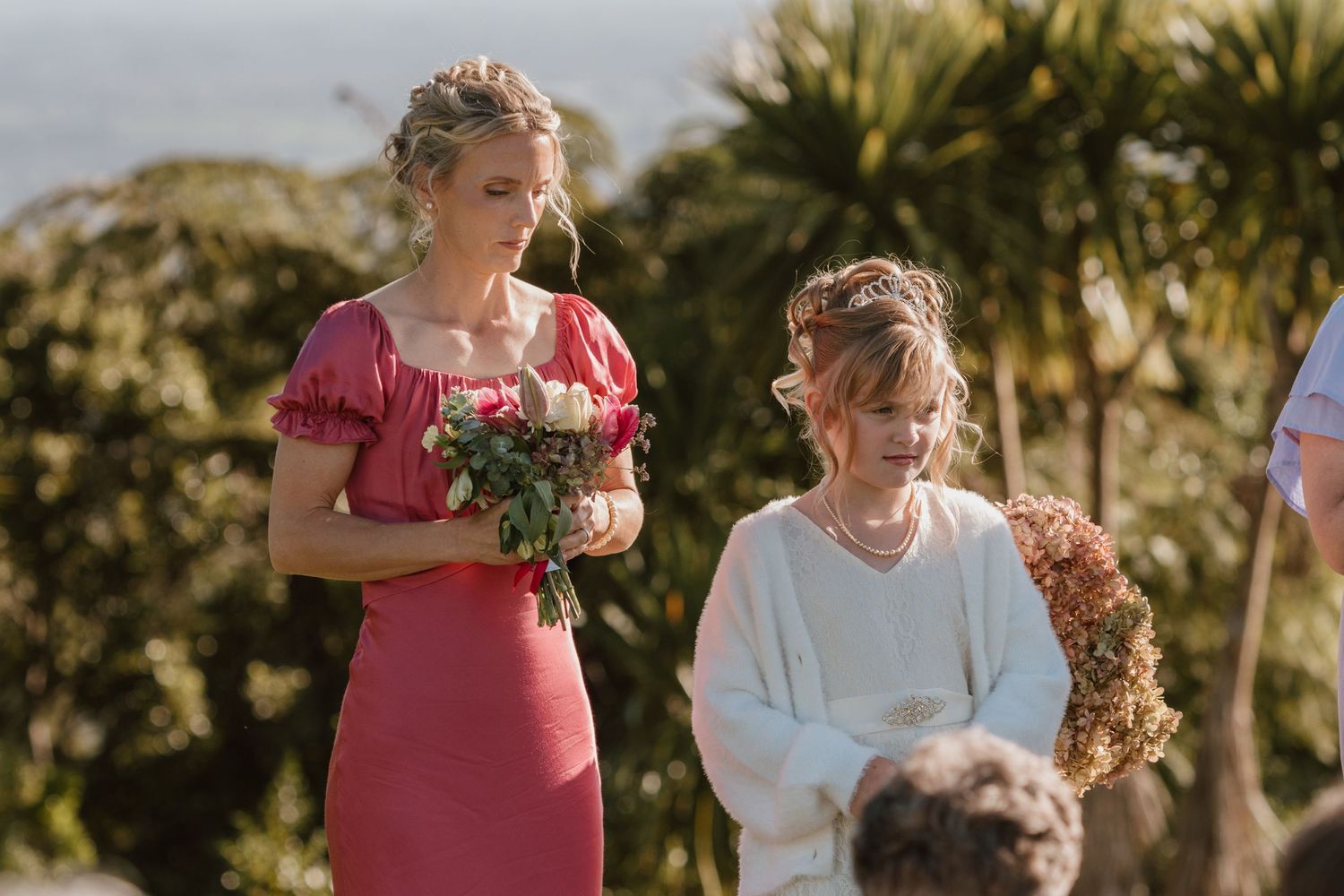 A wedding bridal party stands in an outdoor garden setting during golden hour sunset with palm trees in the background.