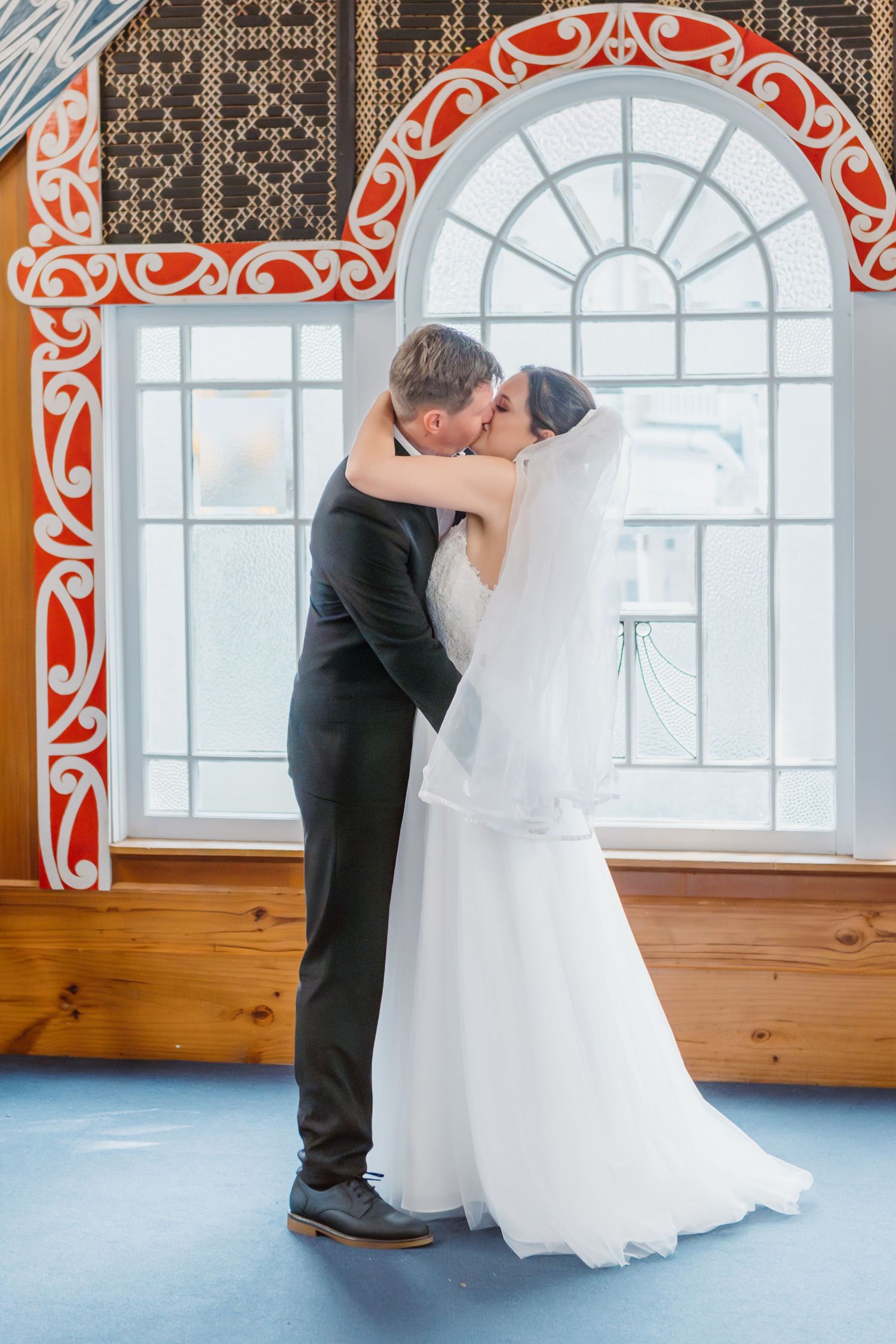 A romantic wedding moment captured in front of an ornate arched window with decorative red trim and wooden floors.
