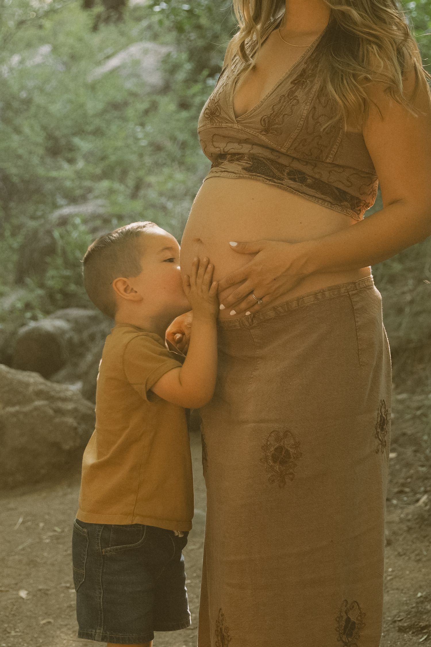 Sweet maternity moment captured in golden sunlight with natural forest backdrop.