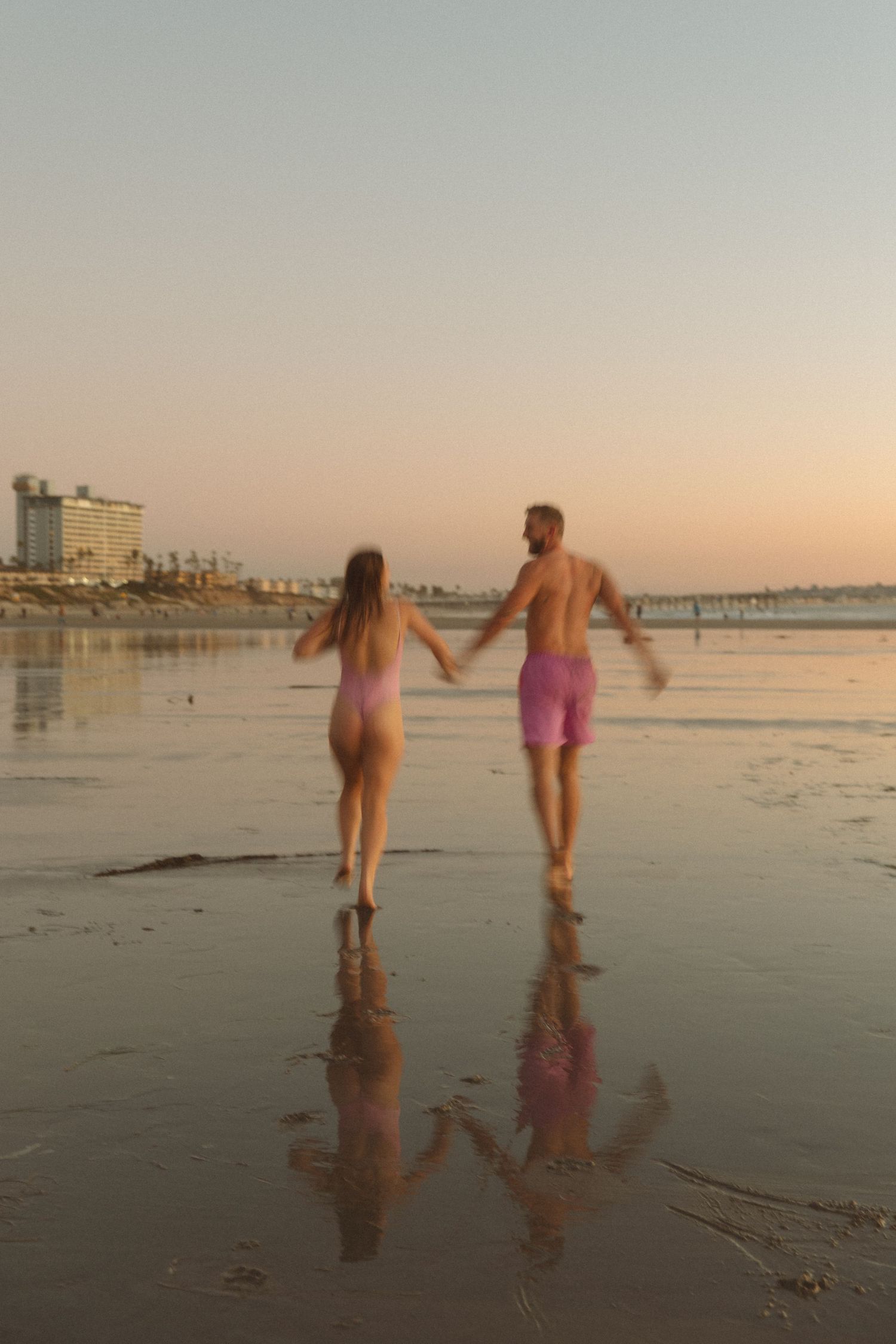 Couple holding hands while walking on a beach at sunset with their reflections in the wet sand.