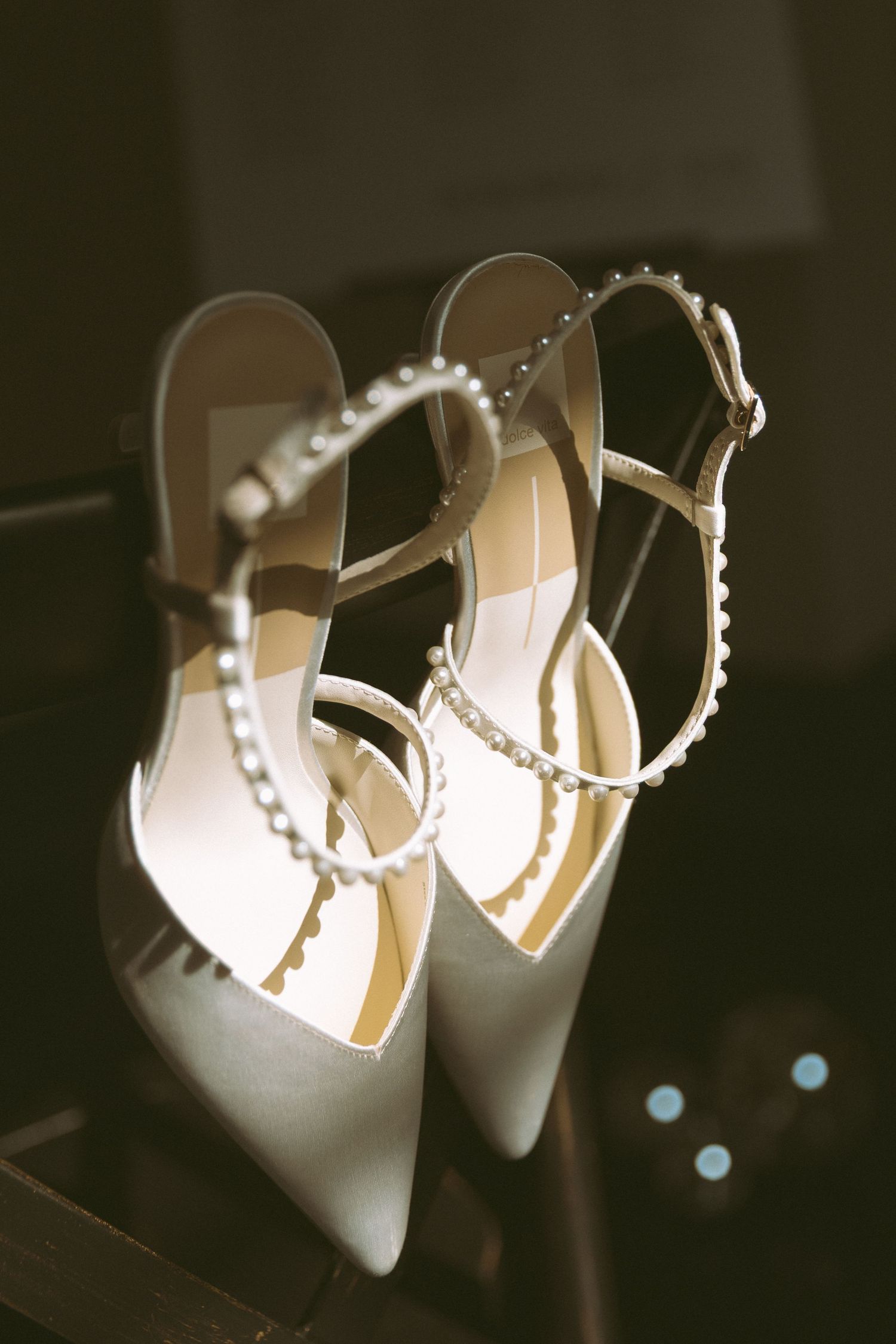 Elegant white satin bridal heels with pearl-embellished ankle straps displayed on a dark surface.