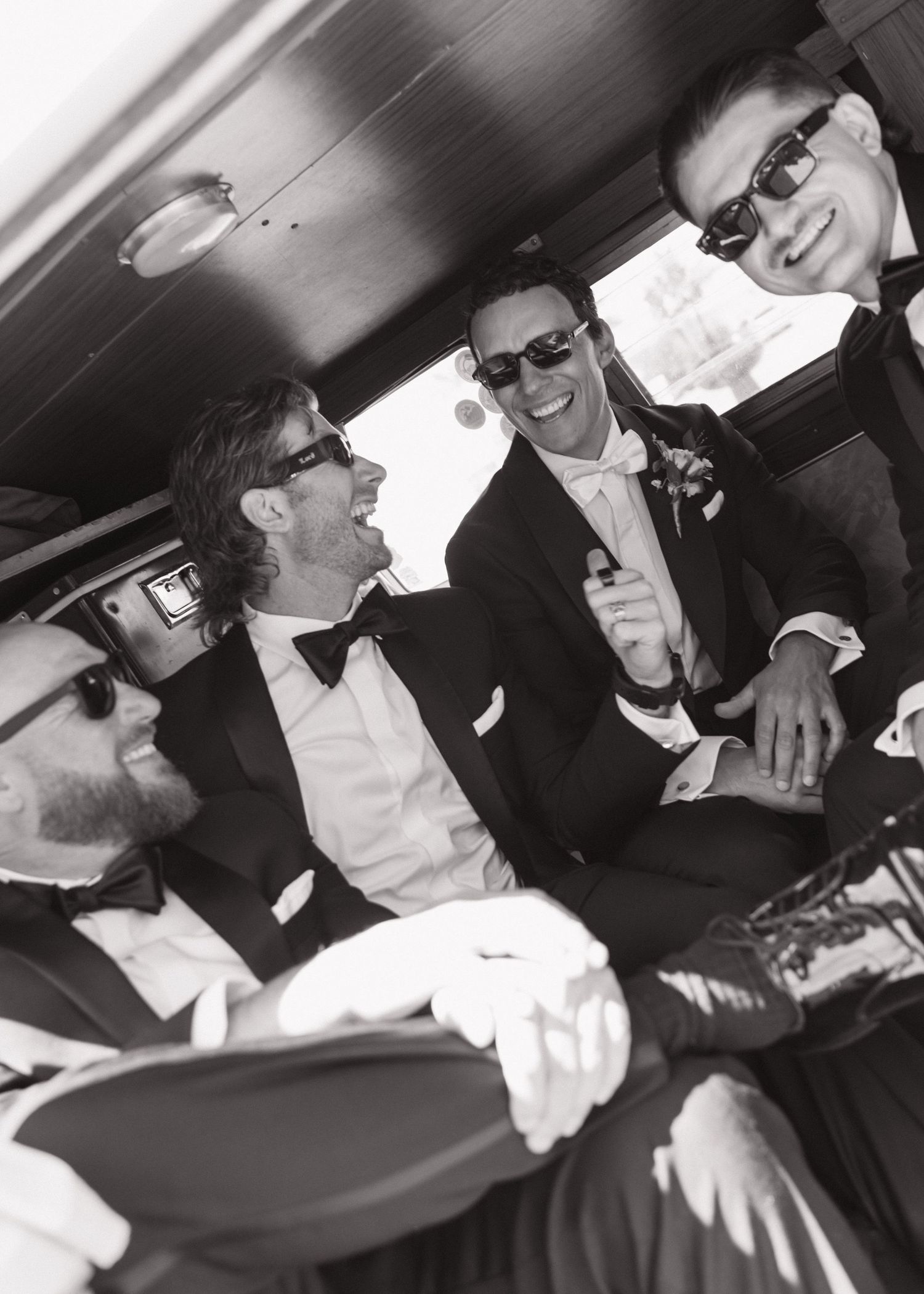 Black and white photo of groom and groomsmen in tuxedos laughing together inside a vintage car.