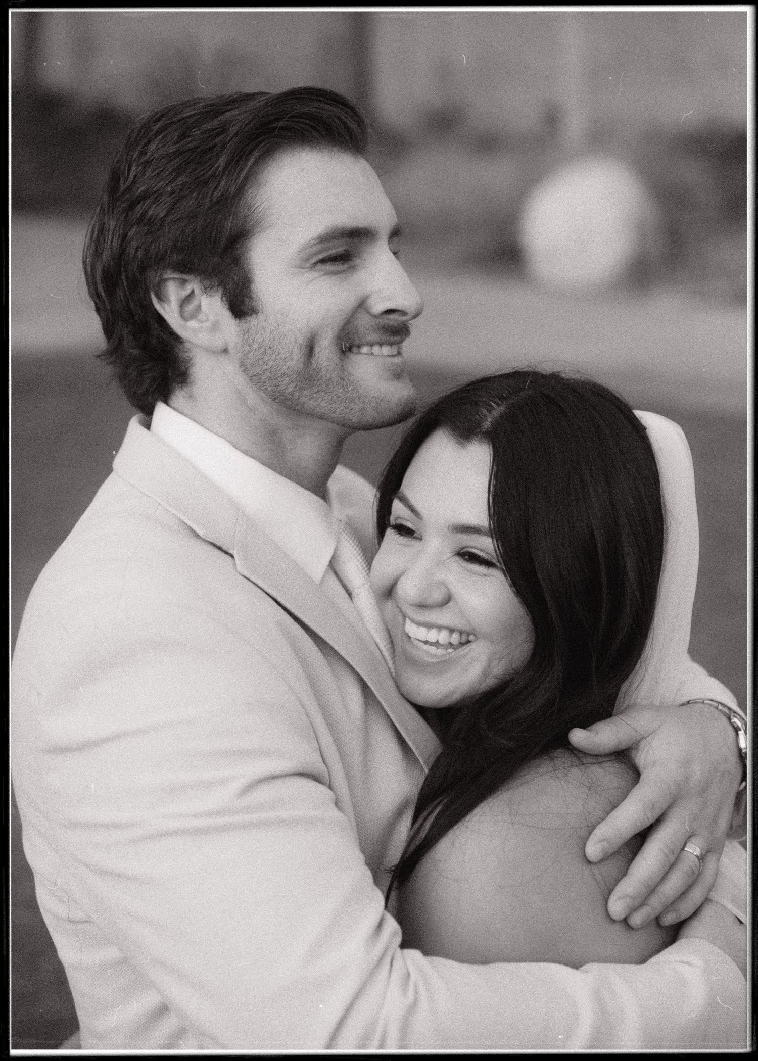 Black and white portrait of a smiling couple embracing, the bride in a veil resting her head on her groom's shoulder, wedding rings visible.
