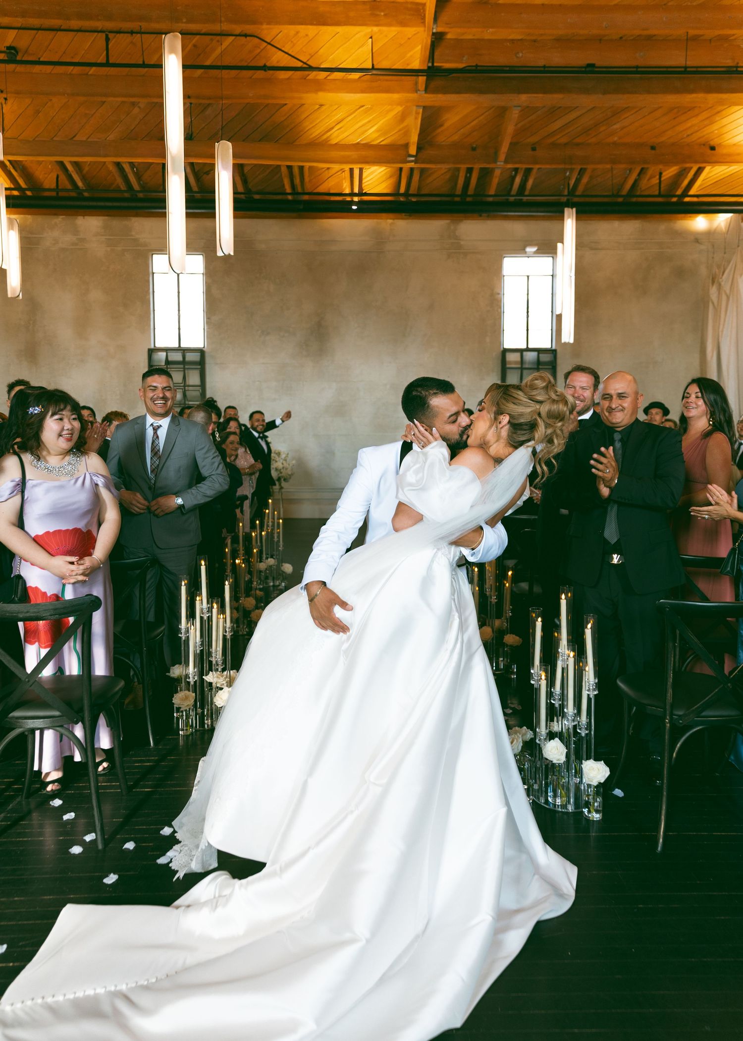 Bride and groom share first kiss at wedding ceremony aisle with guests applauding.