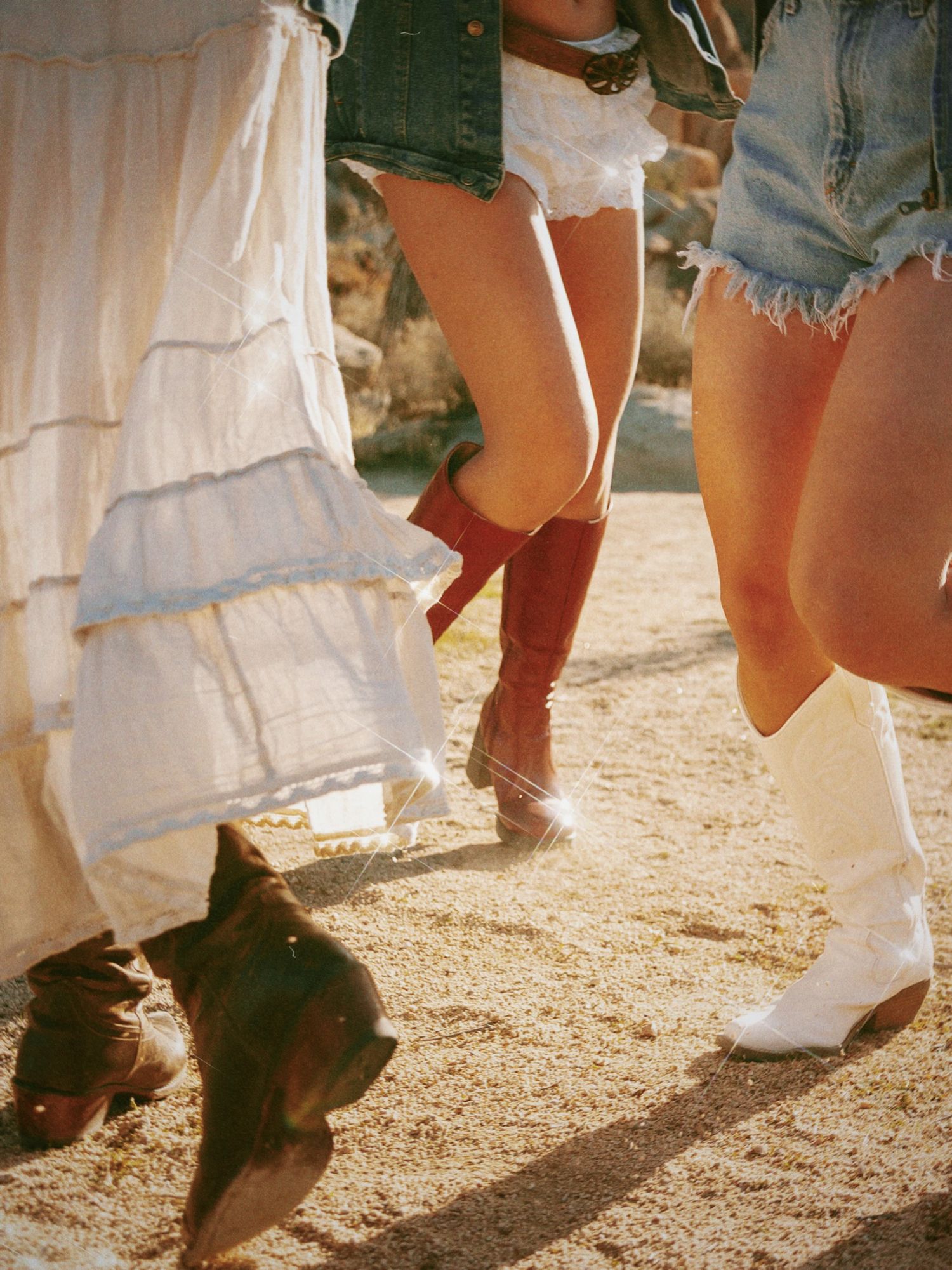 A row of feet wearing cowboy boots and denim shorts standing on dusty ground in bright sunlight.