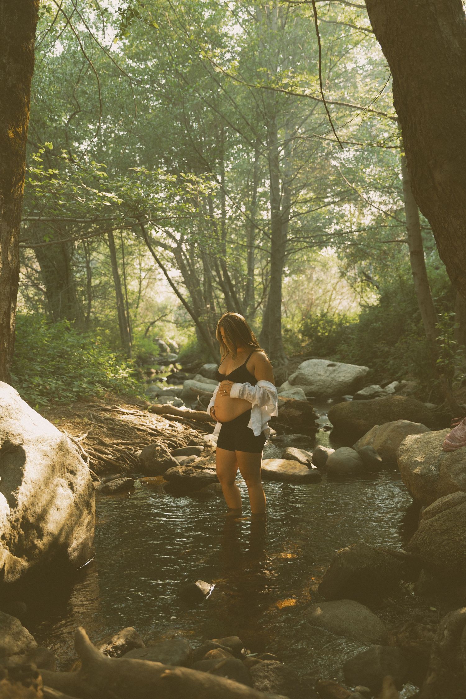 A person in swimwear poses near rocks and water in a wooded setting.