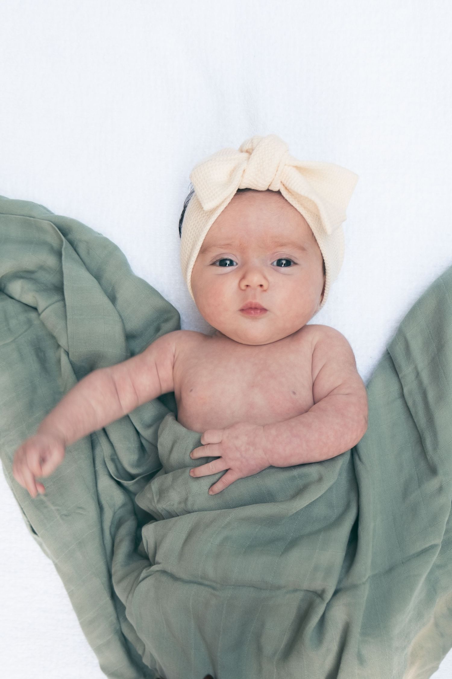 A baby wearing a cream bow headband lies on sage green fabric.