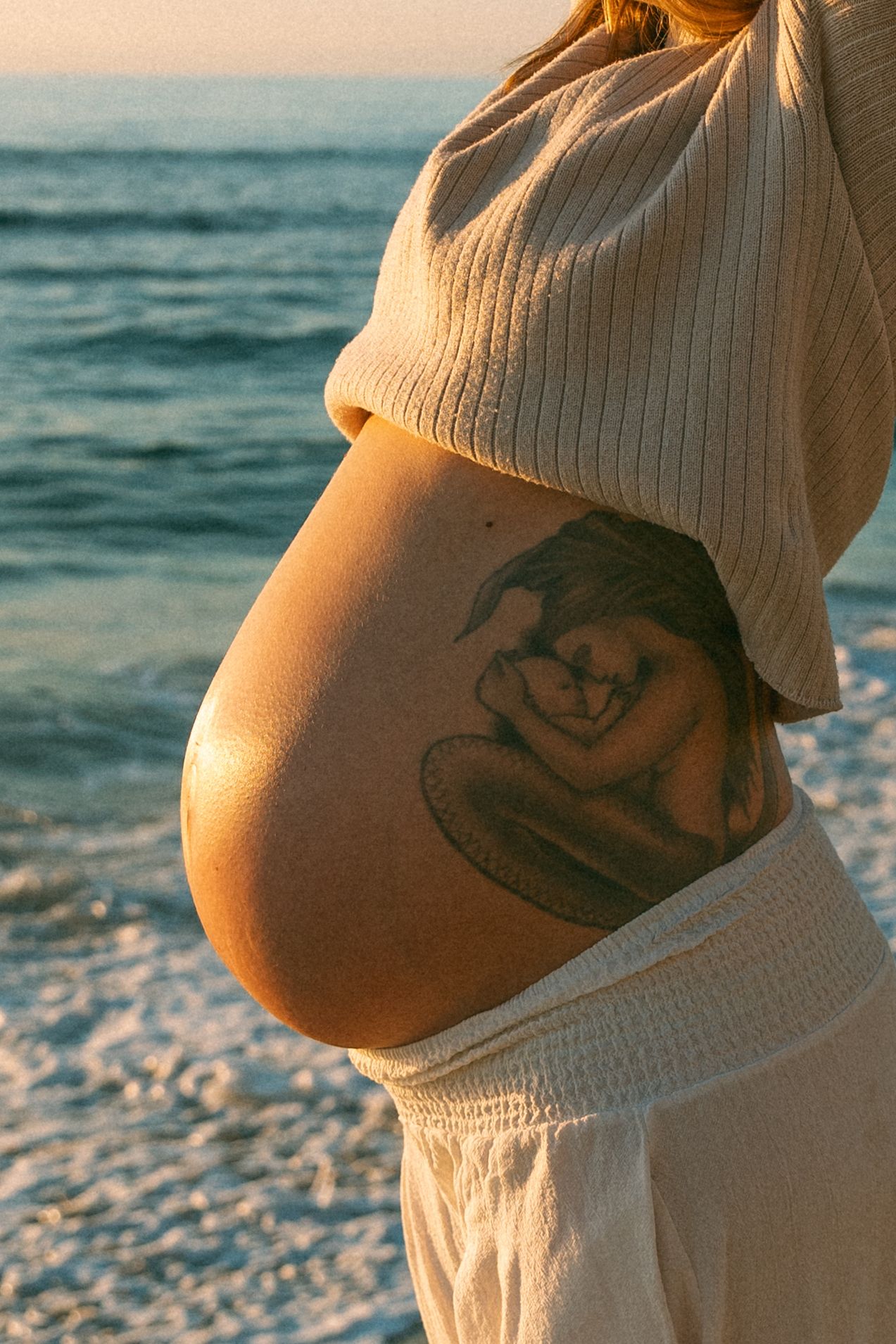 Close-up of pregnant belly with decorative tattoo against ocean background at sunset.