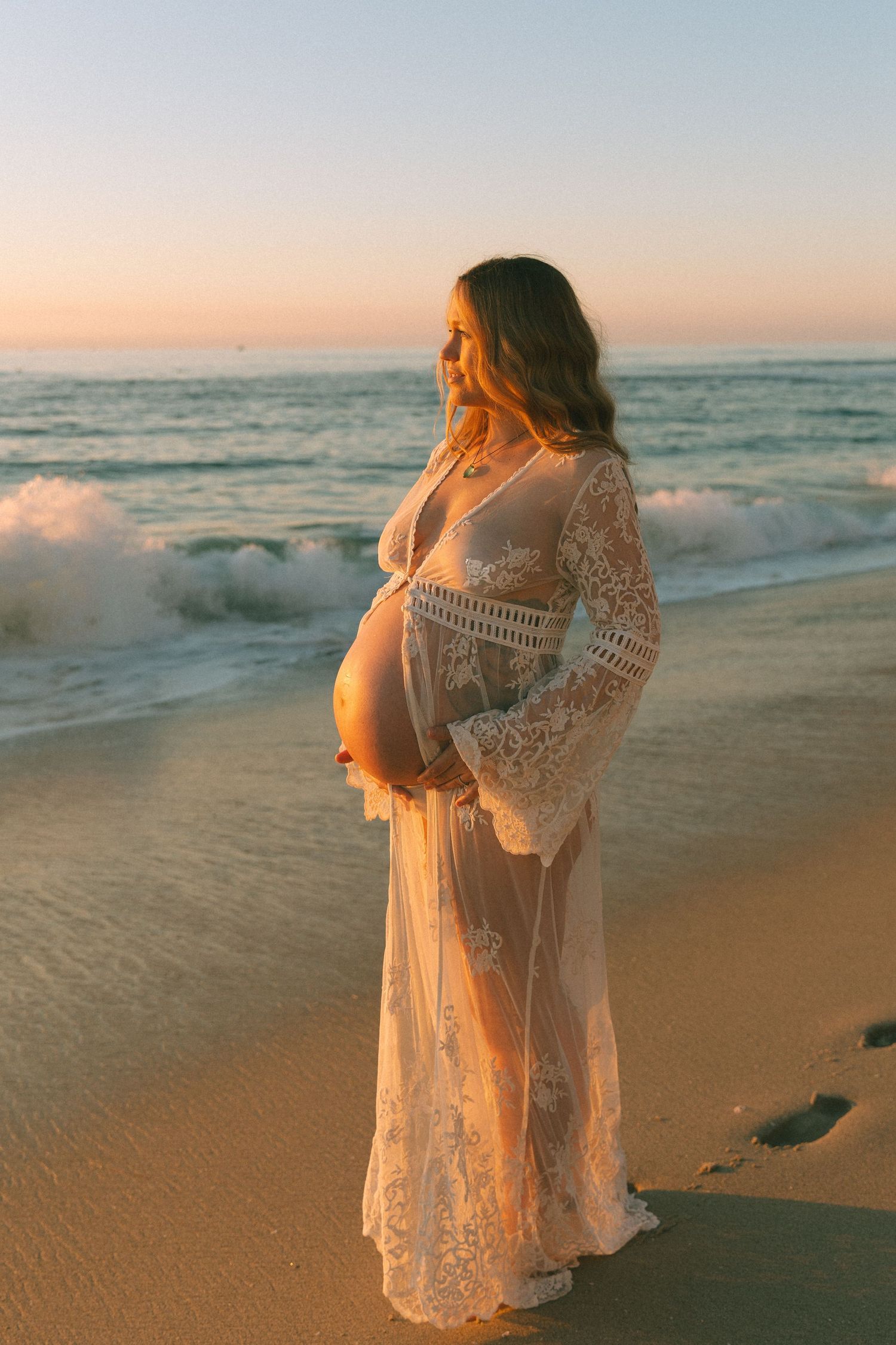 Beachside maternity portrait in white lace dress during golden hour.