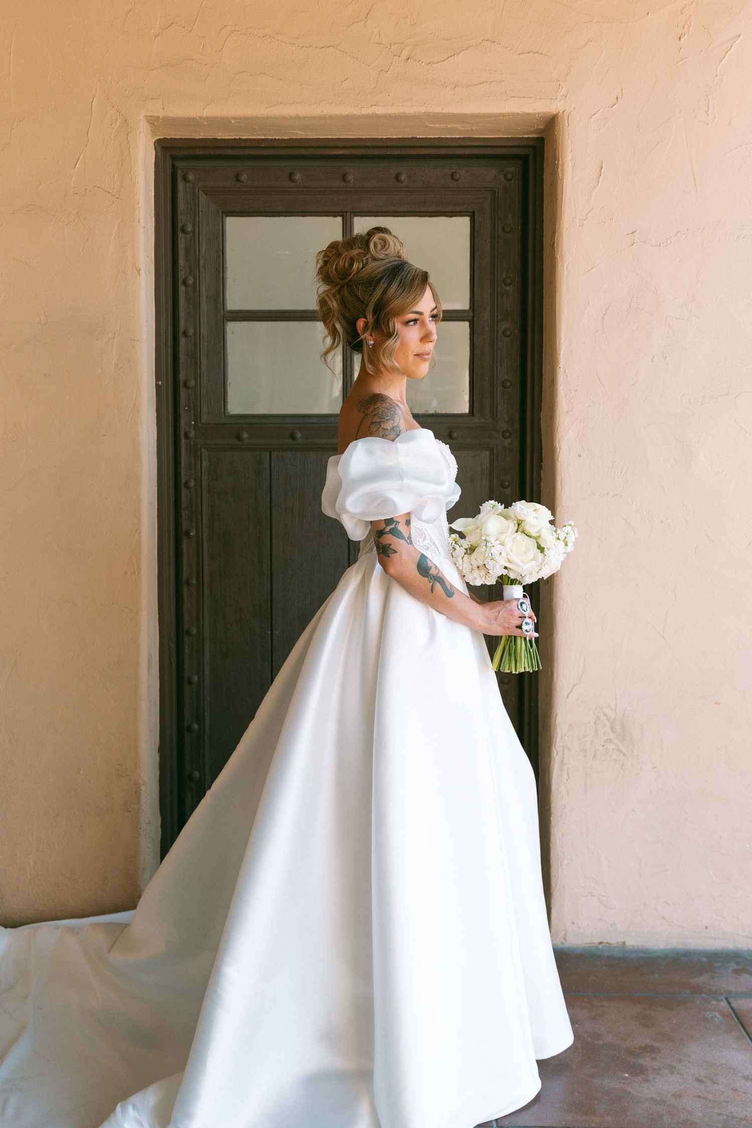 Elegant bride in off-shoulder ball gown holding white bouquet standing by rustic wooden door.
