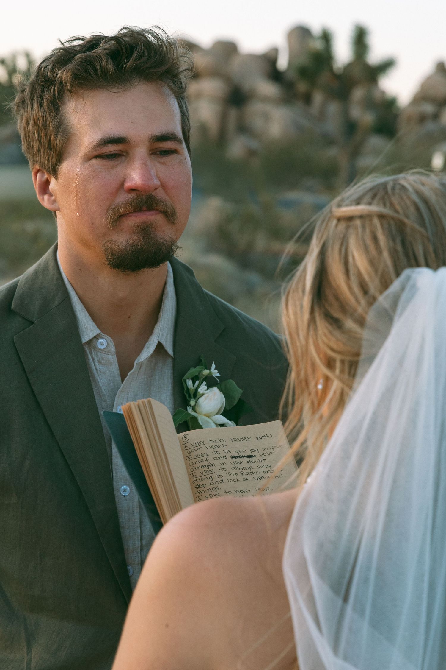 Emotional groom in gray suit with boutonniere listening to bride's vows at outdoor desert wedding.