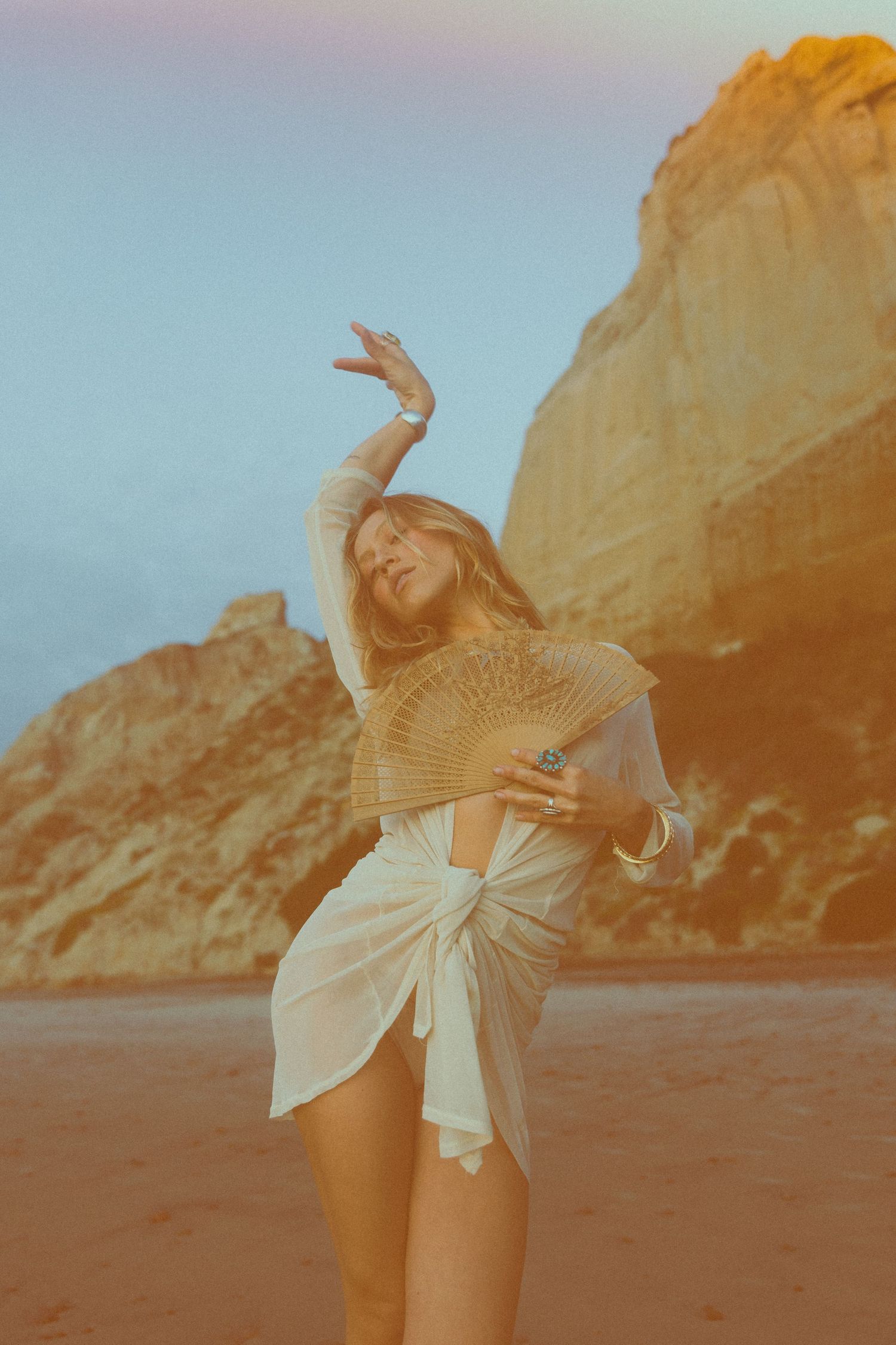A person in a white top dances joyfully on a beach with golden cliffs in the background during sunset.