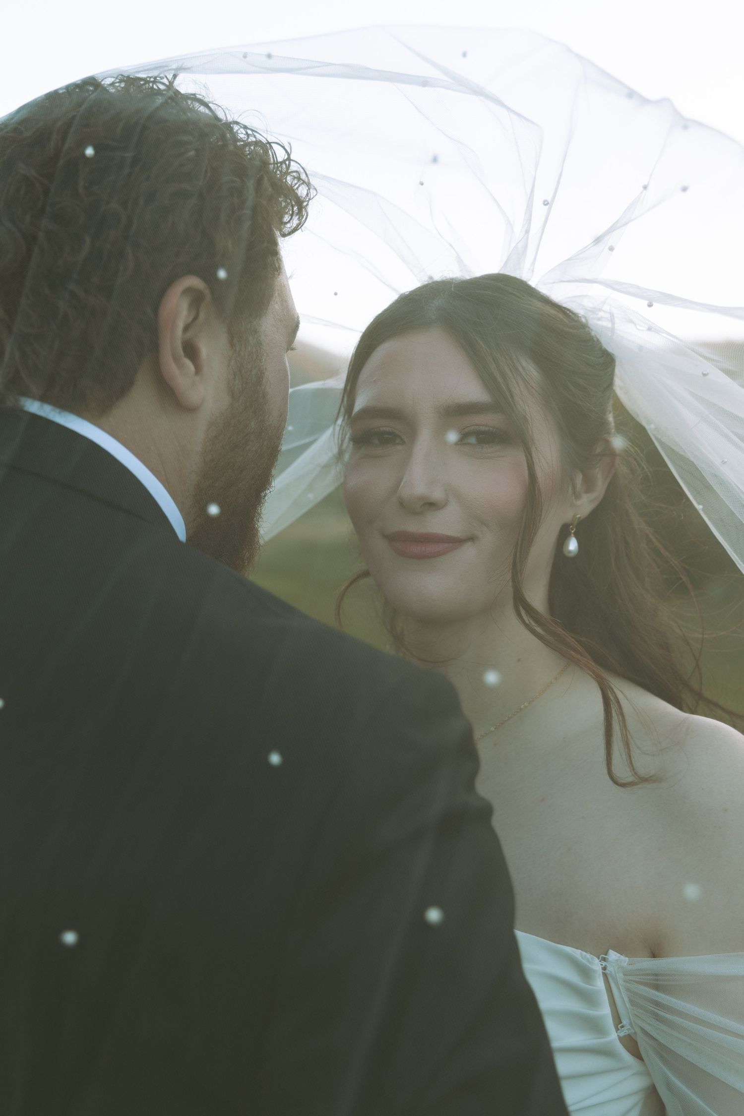 Intimate wedding portrait of couple under flowing veil with soft natural lighting.