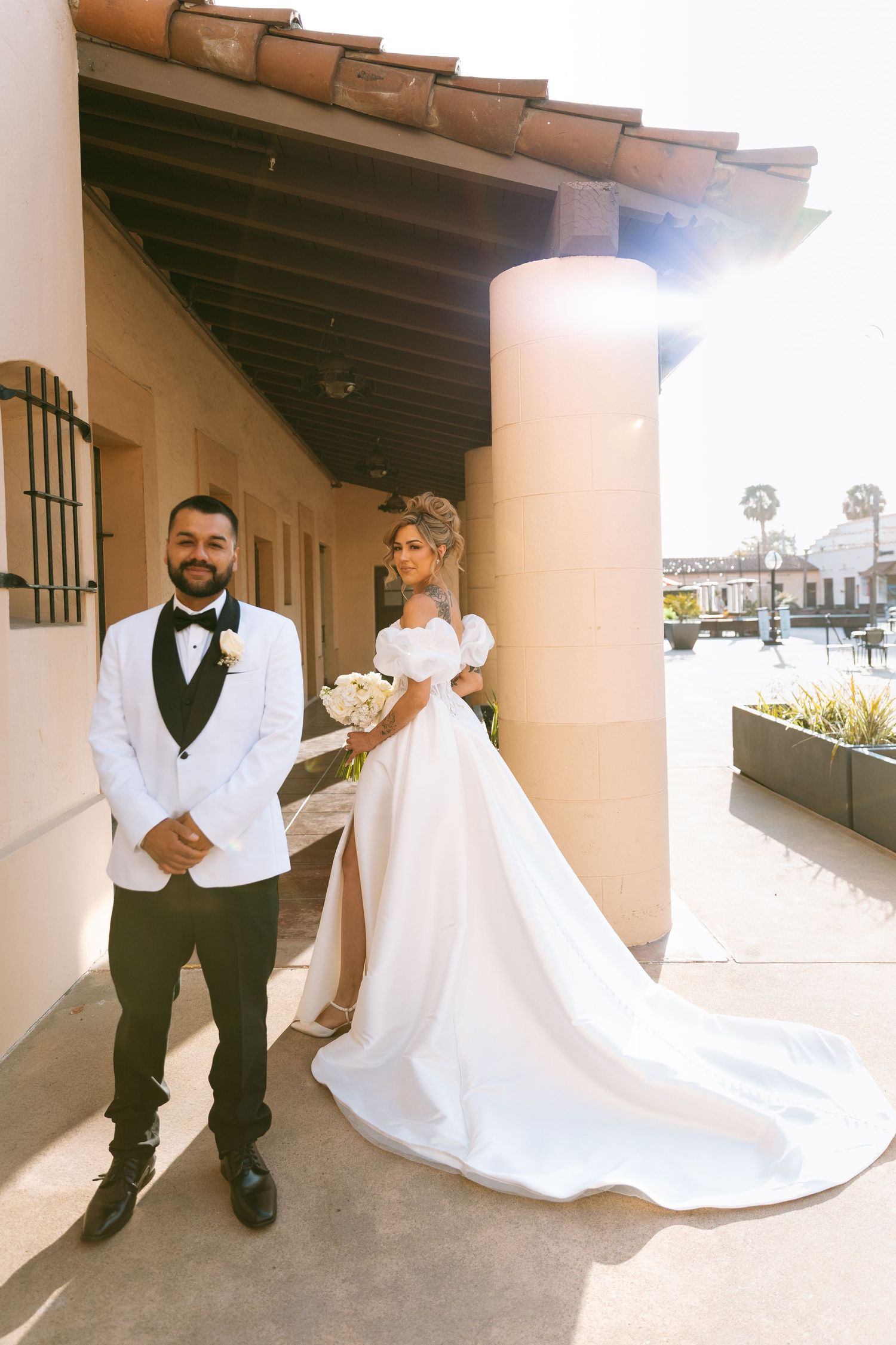 A bride in a white off-shoulder gown with a long train stands beside a groom in a white tuxedo jacket outdoors.