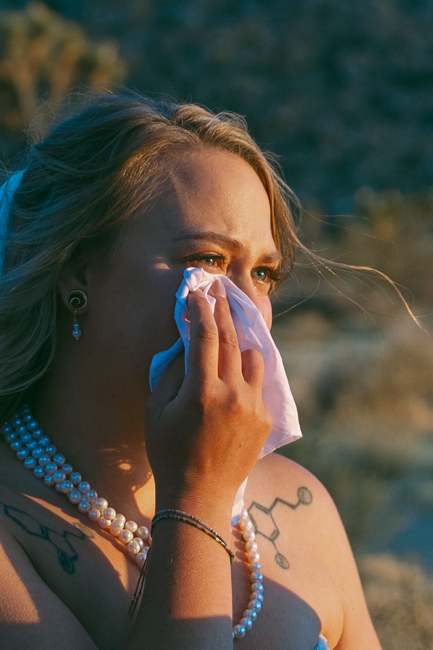 Emotional bride in pearl necklace dabbing tears with tissue outdoors in golden light.