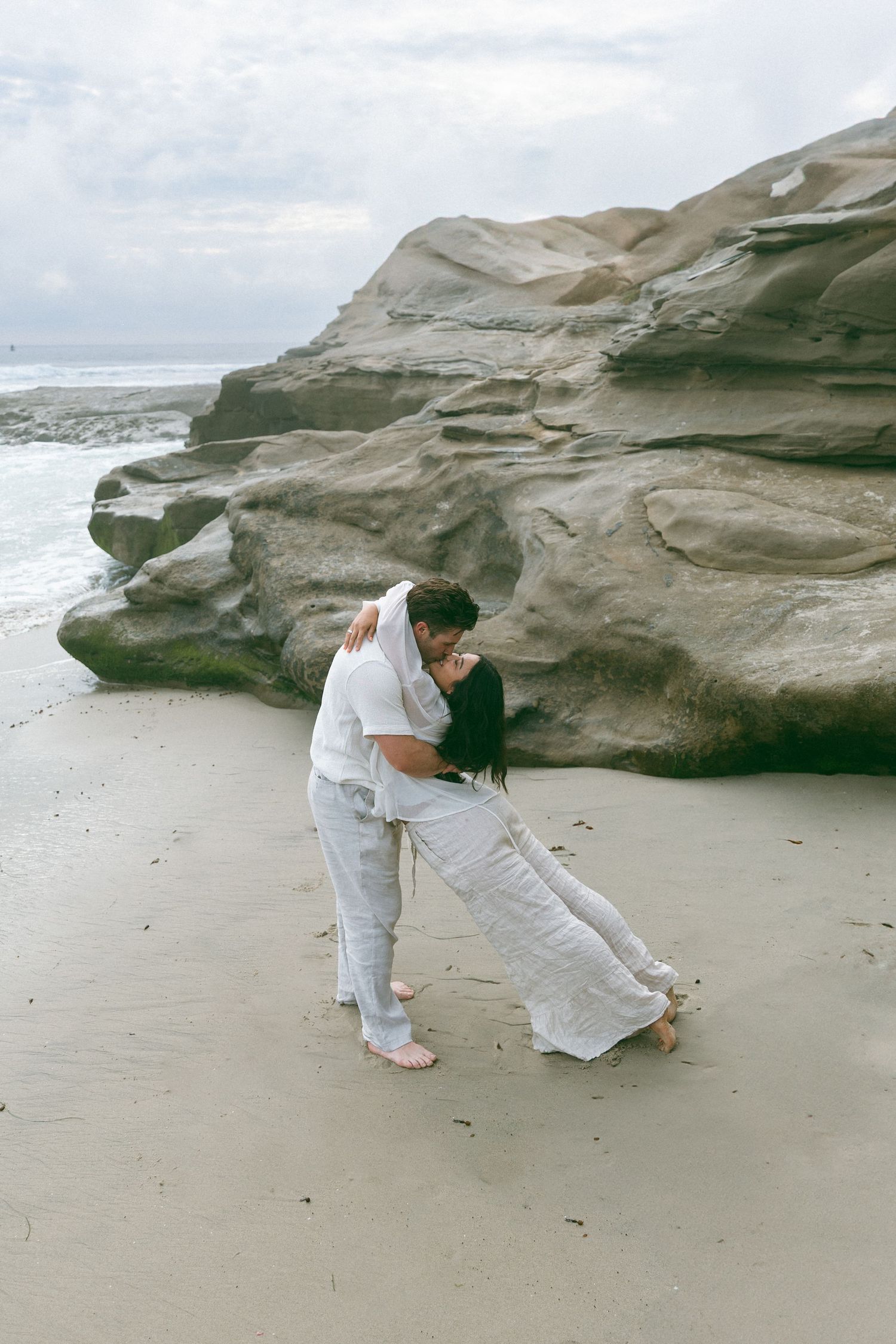 Two people in white outfits pose artistically on a sandy beach with rock formations.