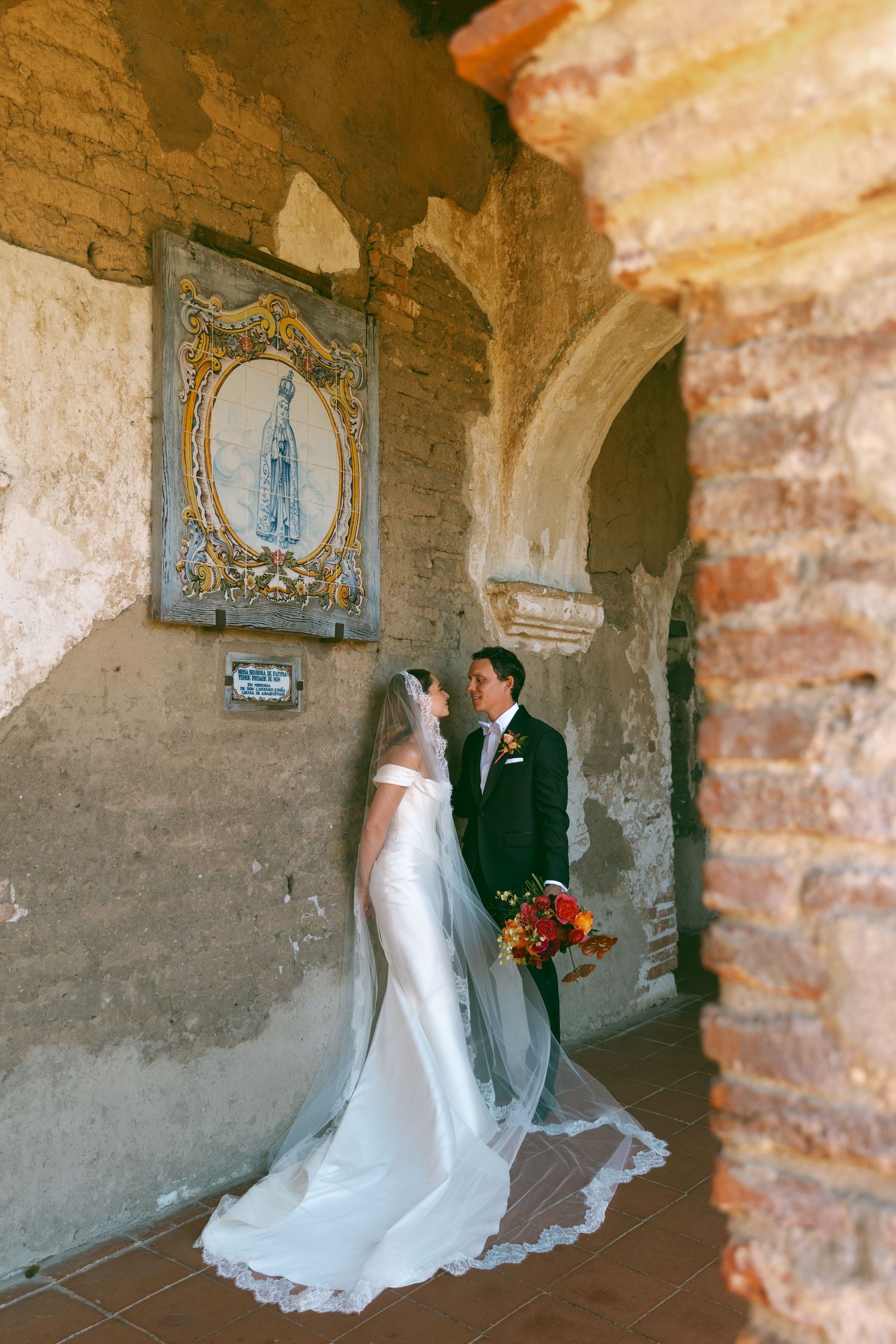 Bride and groom stand together in a rustic mission archway, with a ornate religious tile artwork on the aged stone wall.