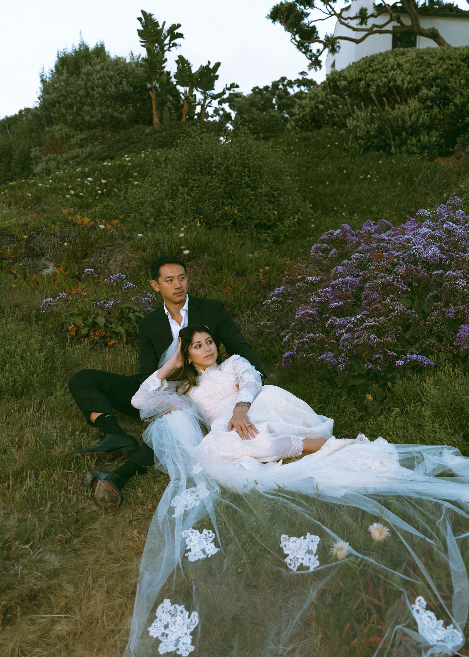 Couple in wedding attire sitting in a wildflower meadow with a lace veil.