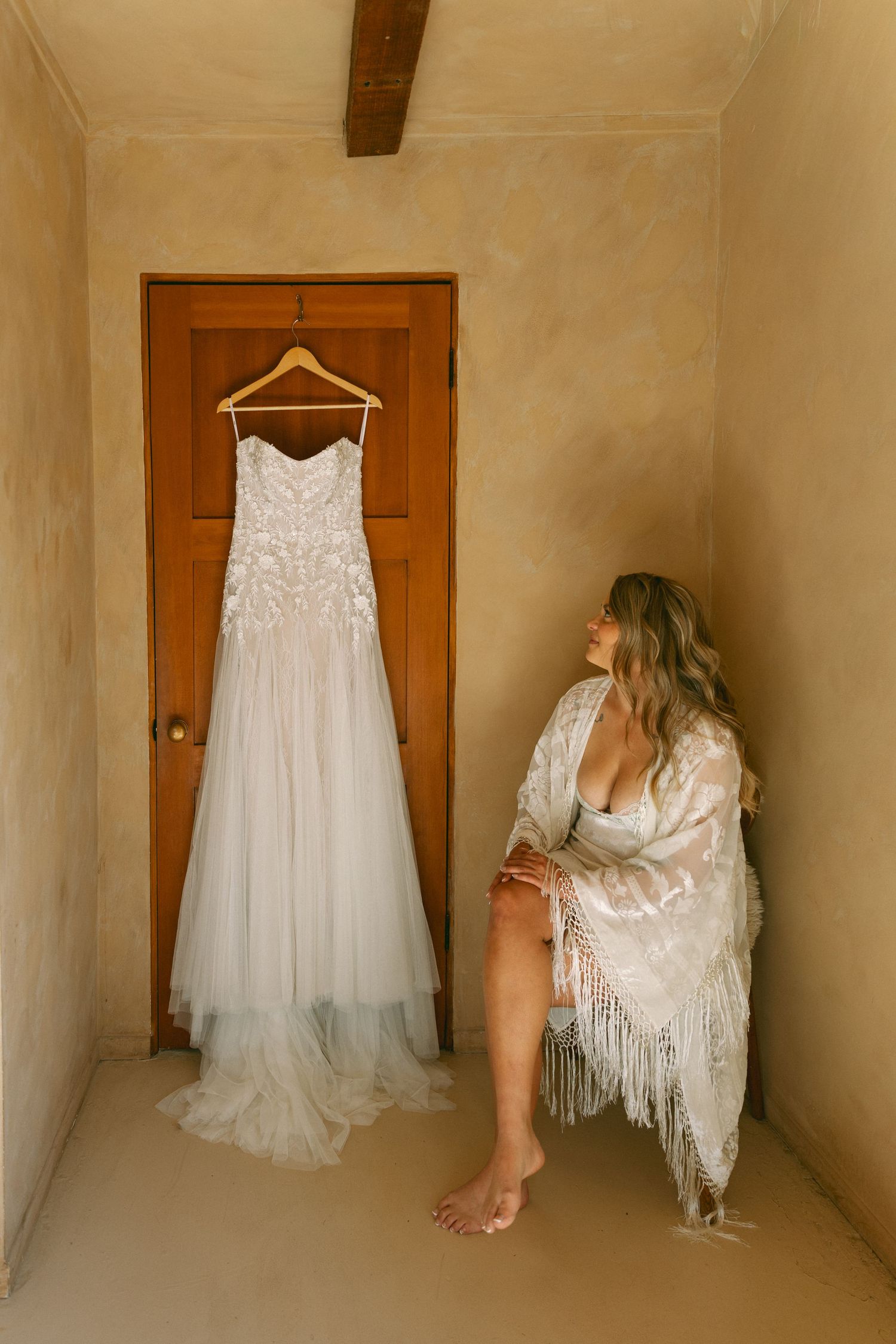 Bride in white lace robe gazes at elegant floral wedding gown hanging on wooden door in rustic adobe room.