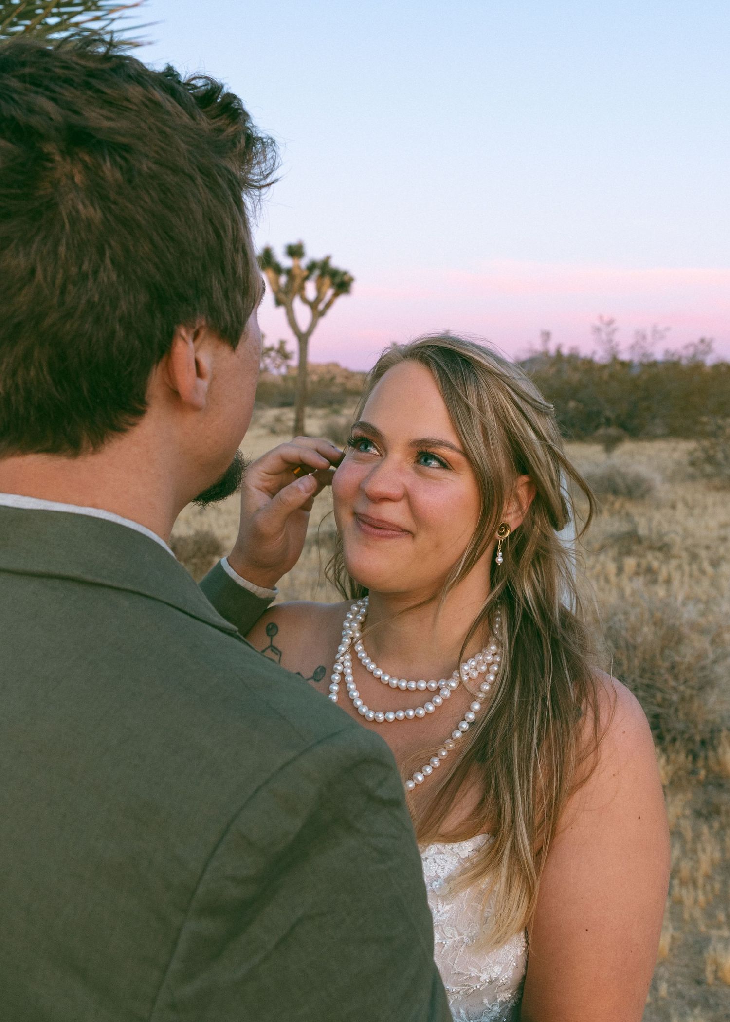 Groom gently wiping bride's tear at desert sunset wedding in Joshua Tree, bride wearing pearl necklace and lace dress.