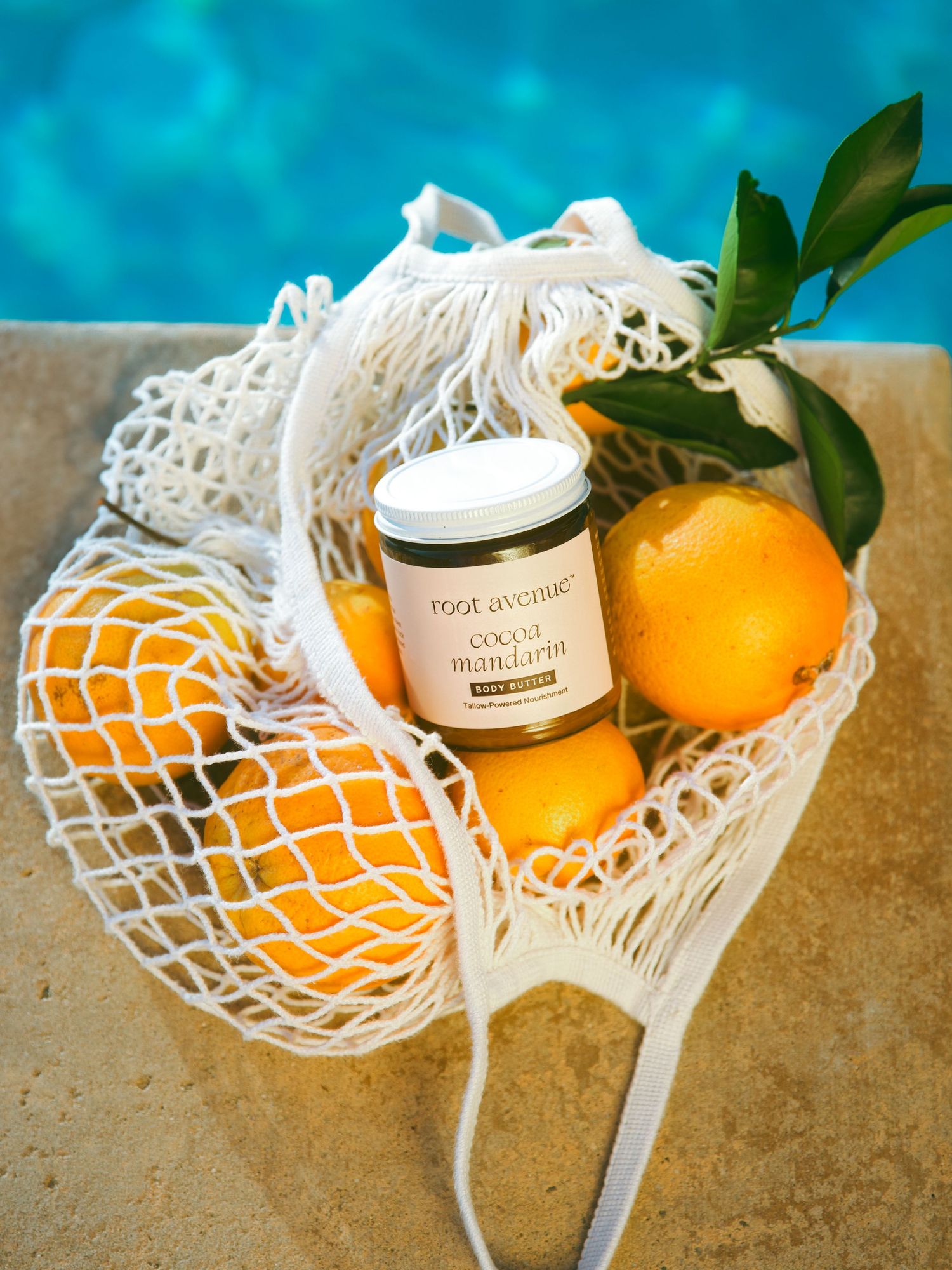 White mesh bag containing oranges and a candle jar displayed by a pool with turquoise water.
