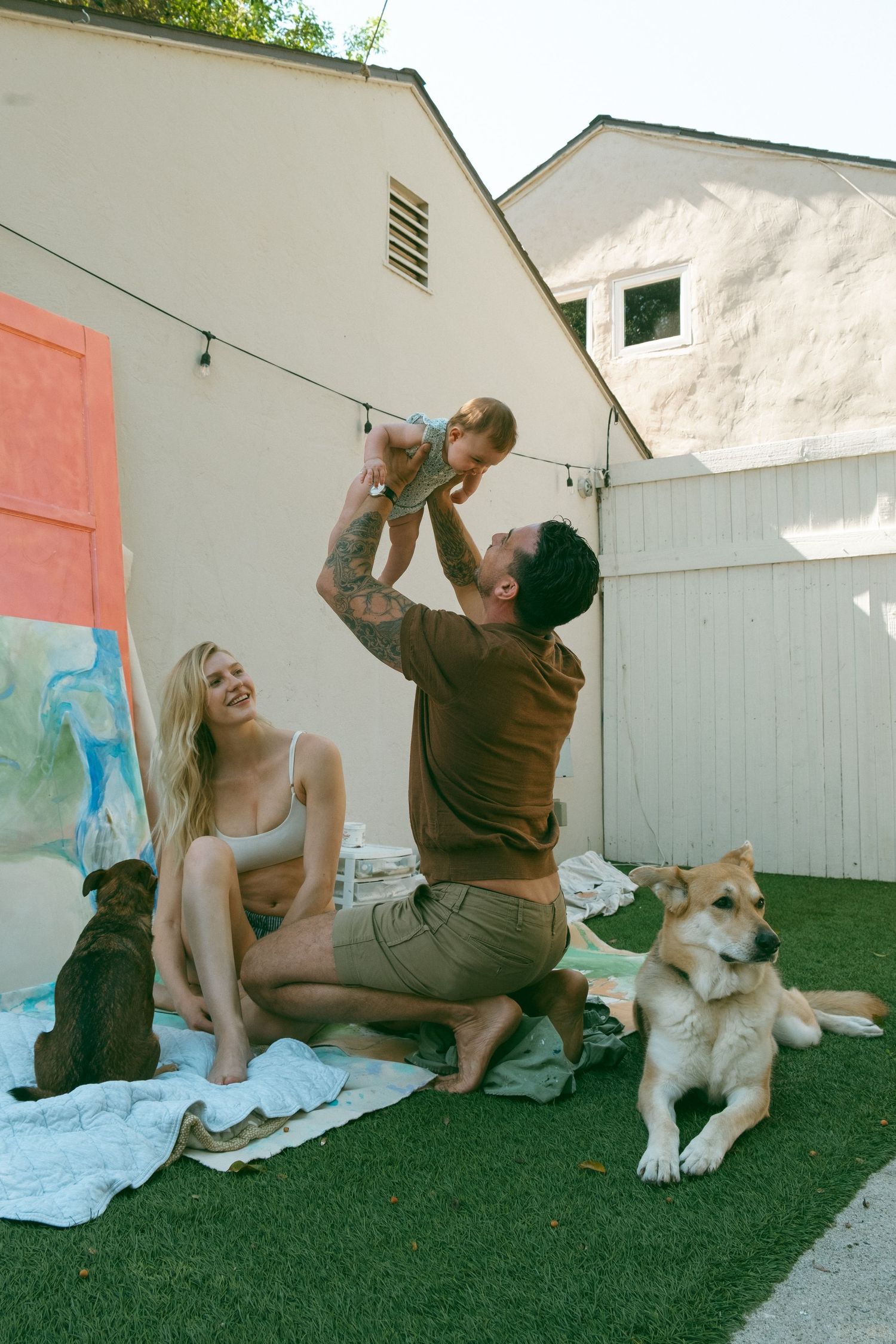 A joyful family scene in a backyard with two dogs and a baby being lifted into the air.