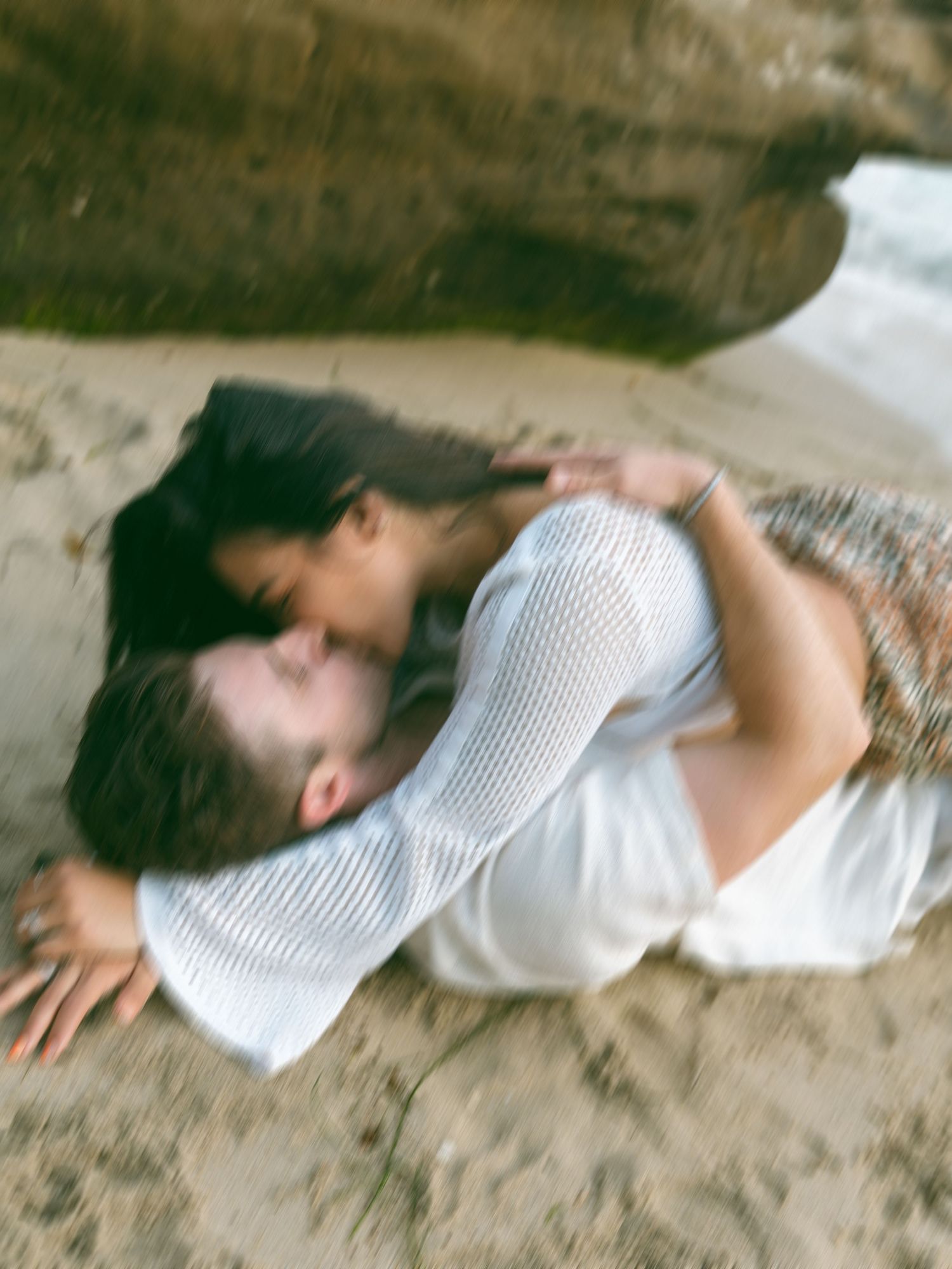 A couple lies together on the sand near coastal rock formations.